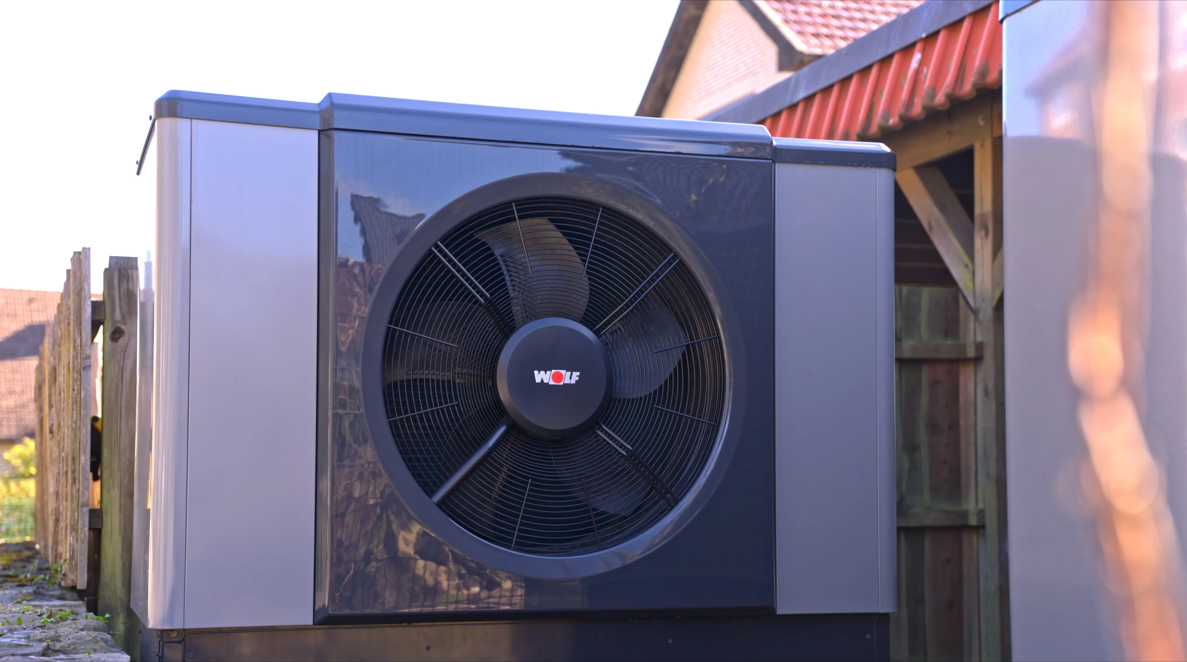 Außenwärmepumpe mit großem Fan und Wolf-Logo, installiert neben einem Zaun und Haus.