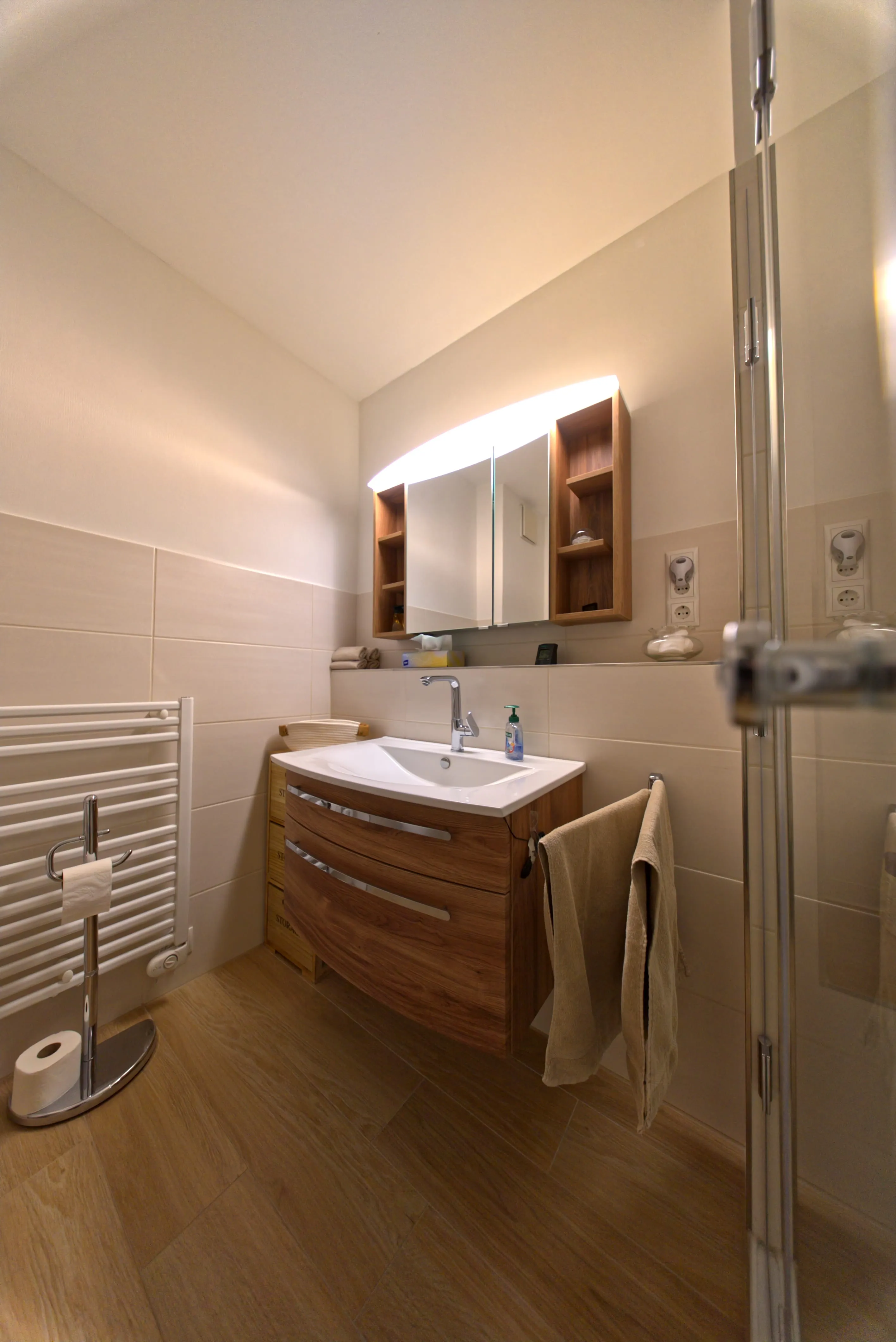 Modernes Badezimmer mit beleuchtetem Spiegel, Holzwaschtisch, Handtuch und WC-Papierhalter.