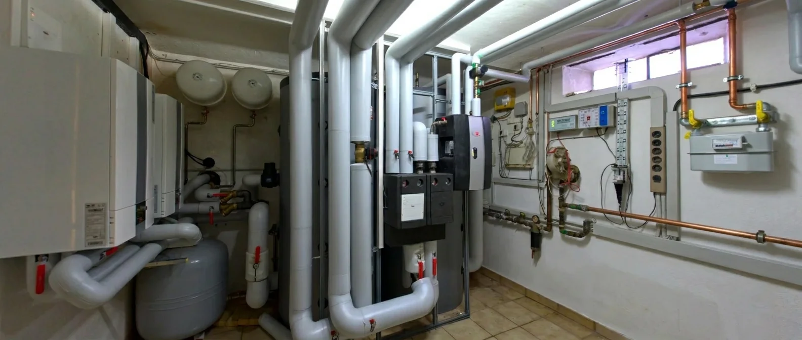 Heizungsanlage mit großen grauen Rohren, Zählern und Kupferleitungen in einem Kellerraum.