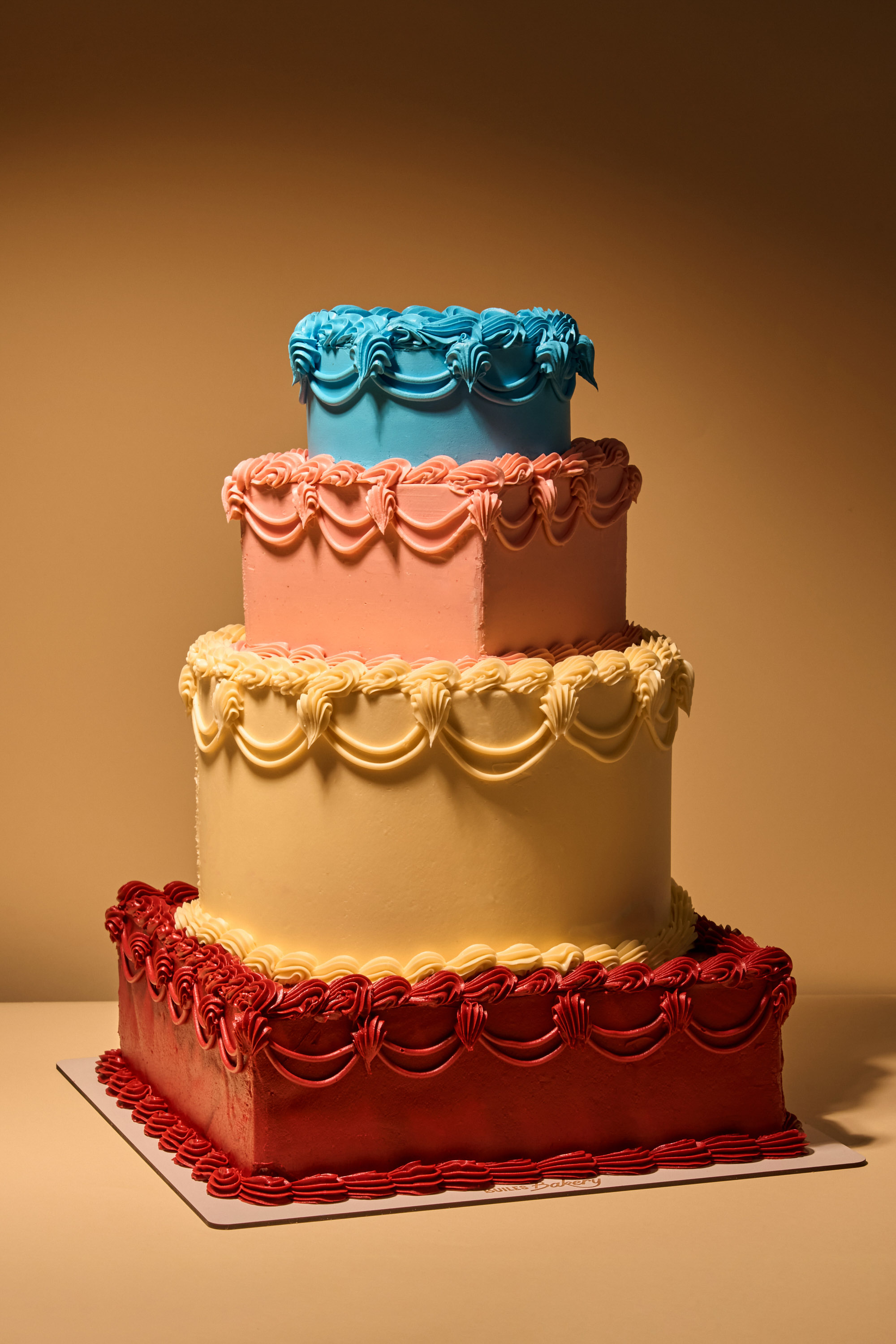 Product photography by Gooper for Builes Bakery, featuring a colorful tiered cake with red, yellow, peach, and blue buttercream layers against a warm studio background.