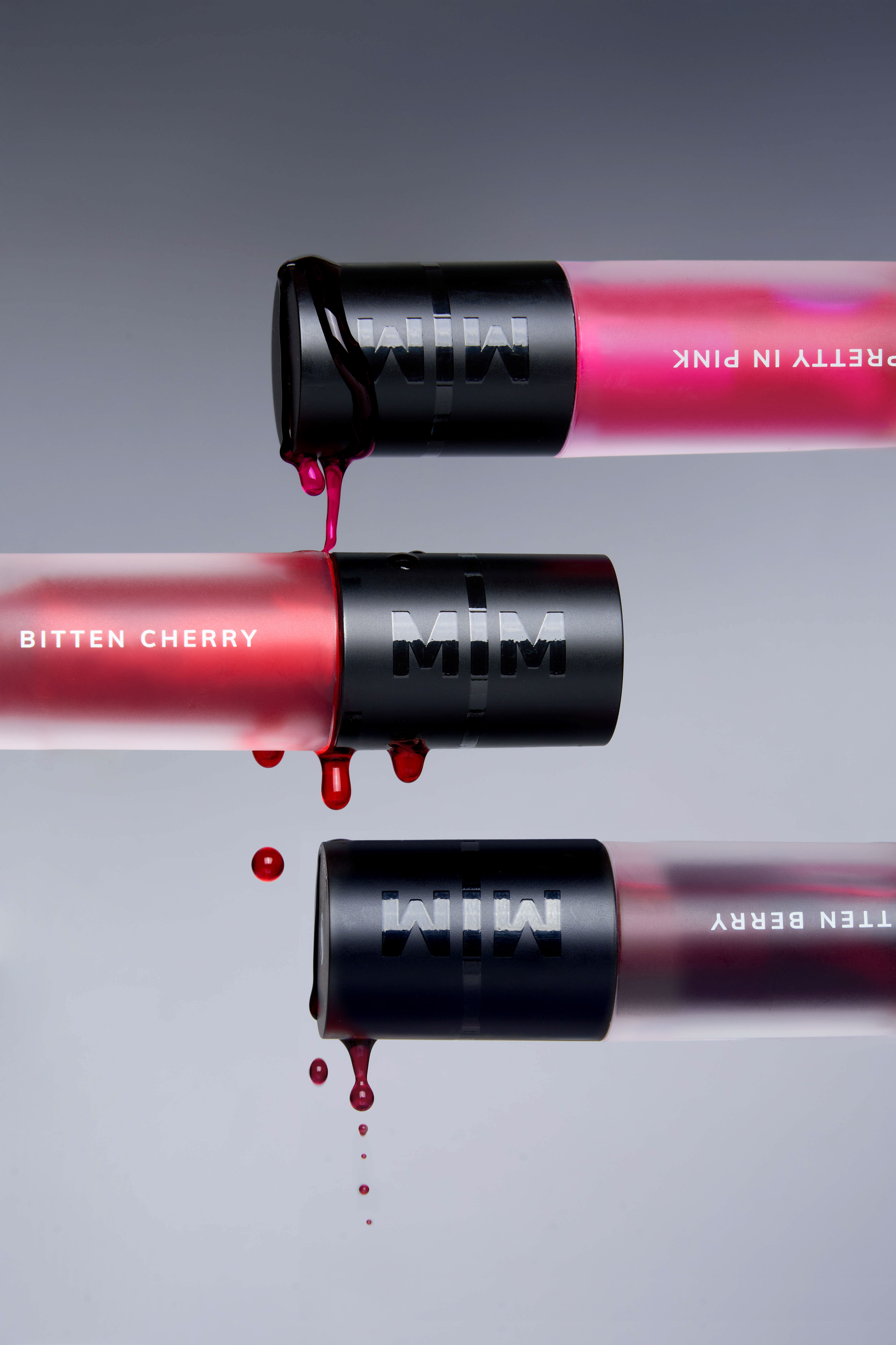 Inverted product photography by Gooper: MIM Pretty in Pink, Bitten Cherry and Bitten Berry lip tints dripping over a gray gradient.