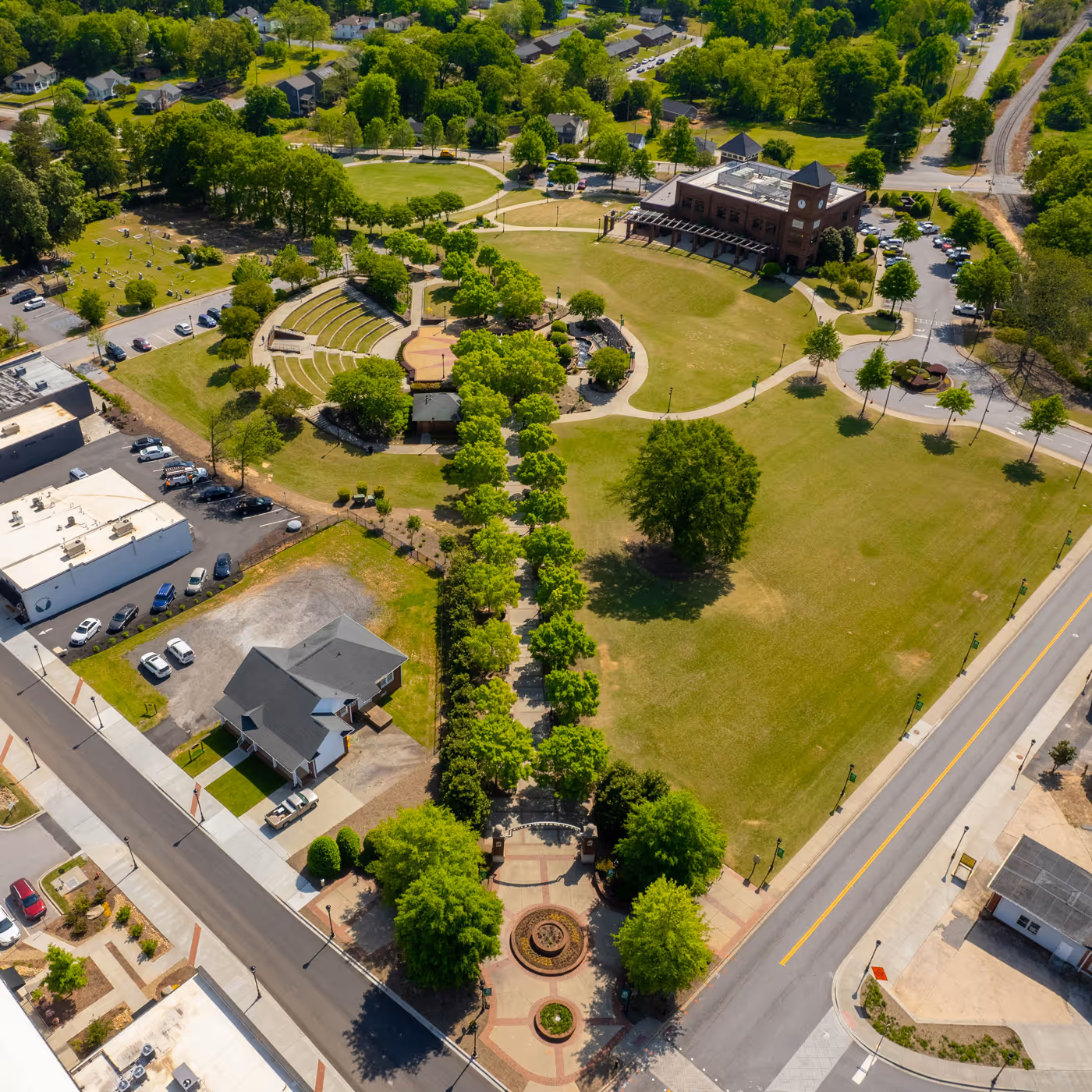Aerial view of Greer's green park with trees, walking paths, an amphitheater, and nearby houses in a suburban area.