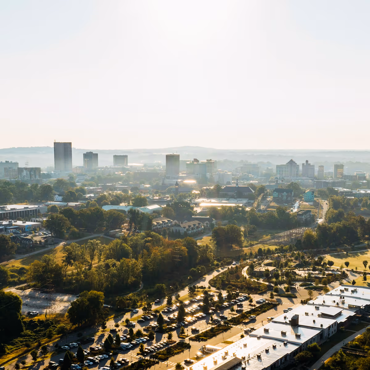 Aerial view of the Greenville skyline with tall buildings, green trees, parking lots, and winding roads under bright daylight.