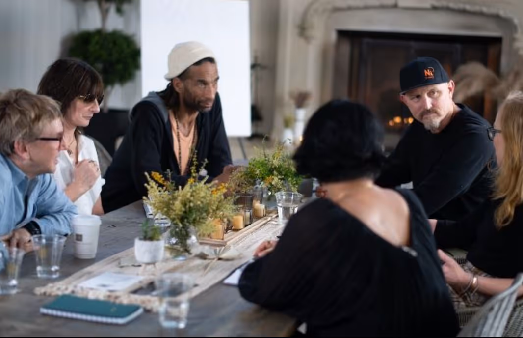 Group of people seated around a table having a focused discussion, listening and speaking in a collaborative indoor setting.