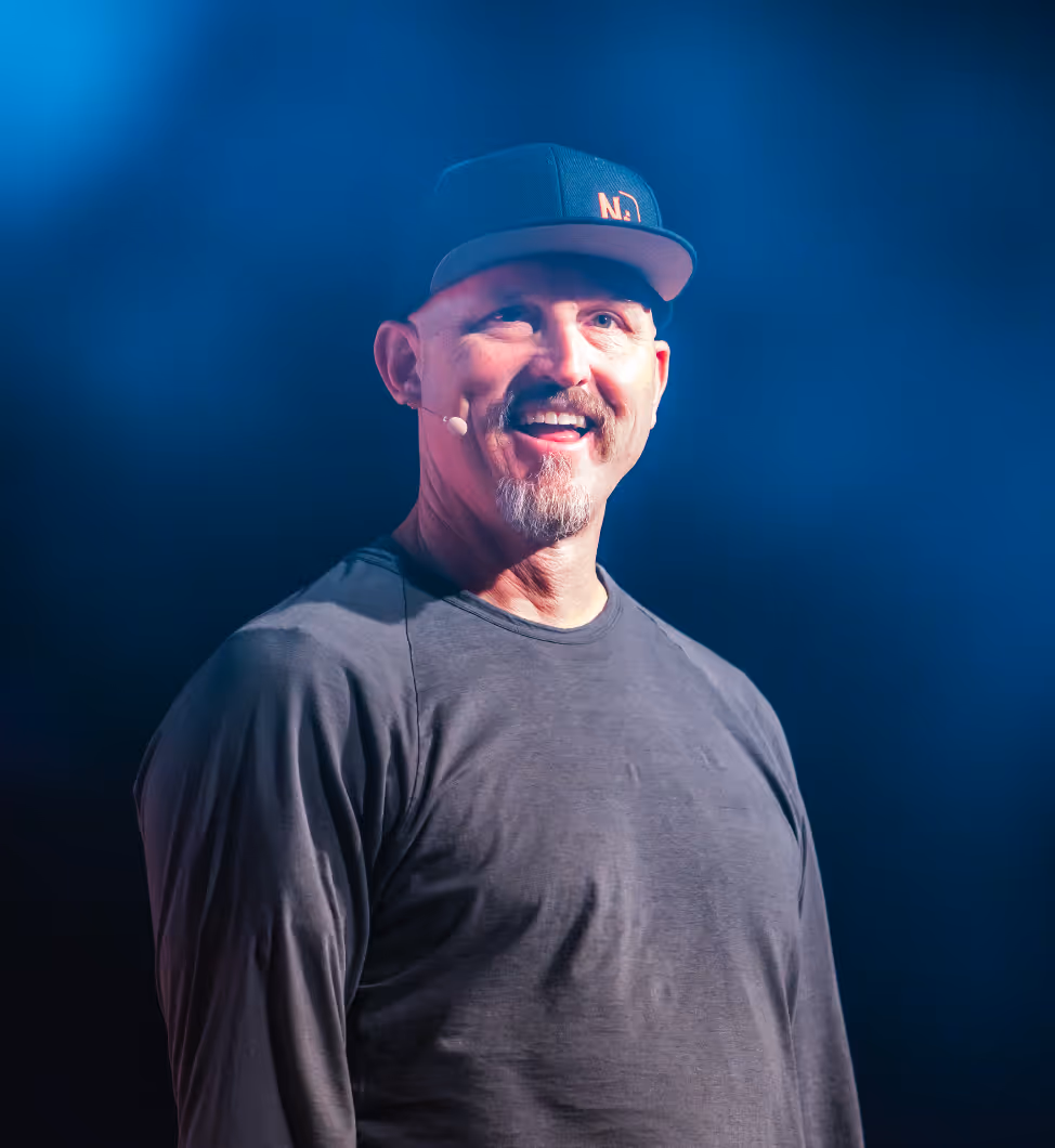 Mick Ebeling speaking on stage, wearing a black cap and headset microphone under blue lighting.