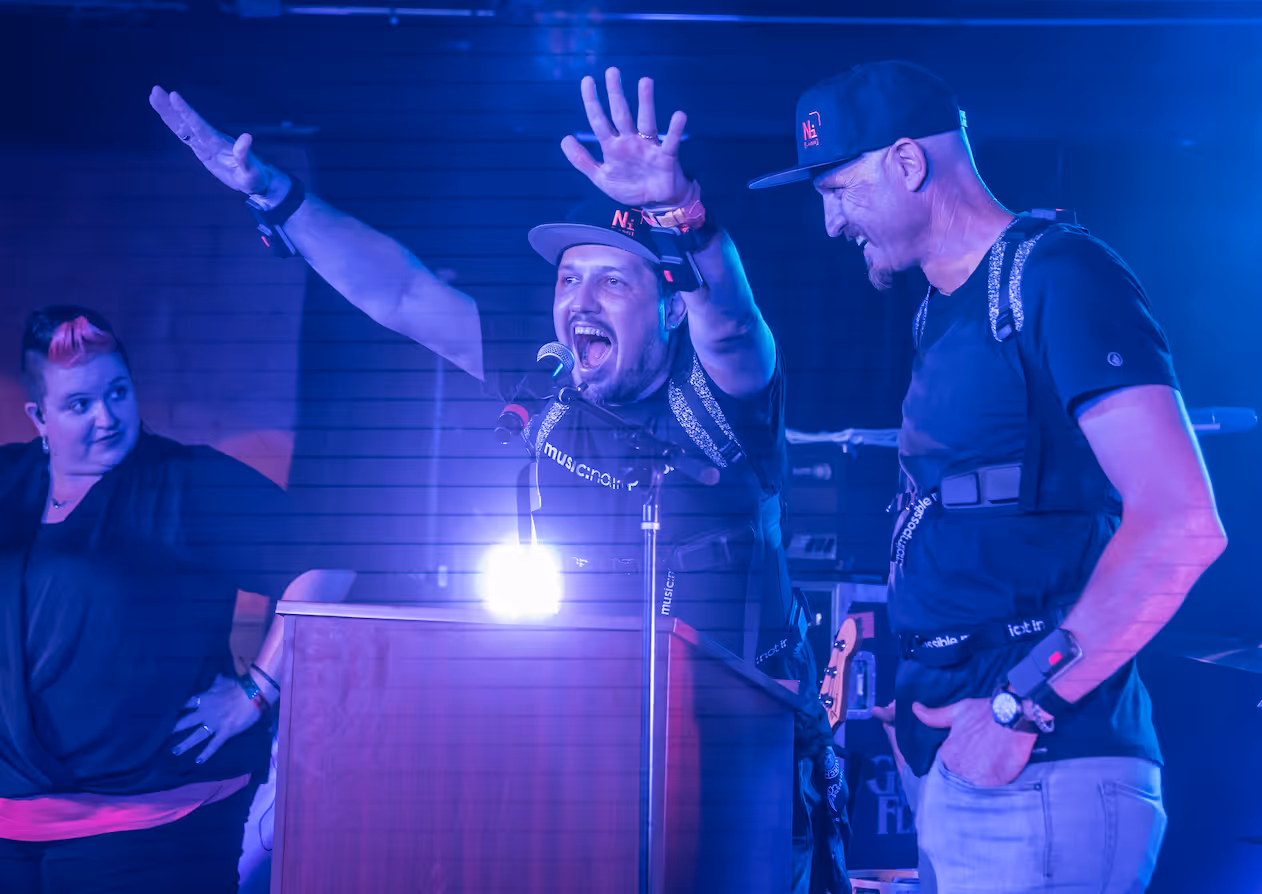 Two inventors of the Not Impossible adaptive music suits stand on stage wearing assistive wearable technology, raising their hands and engaging excitedly with a live audience.