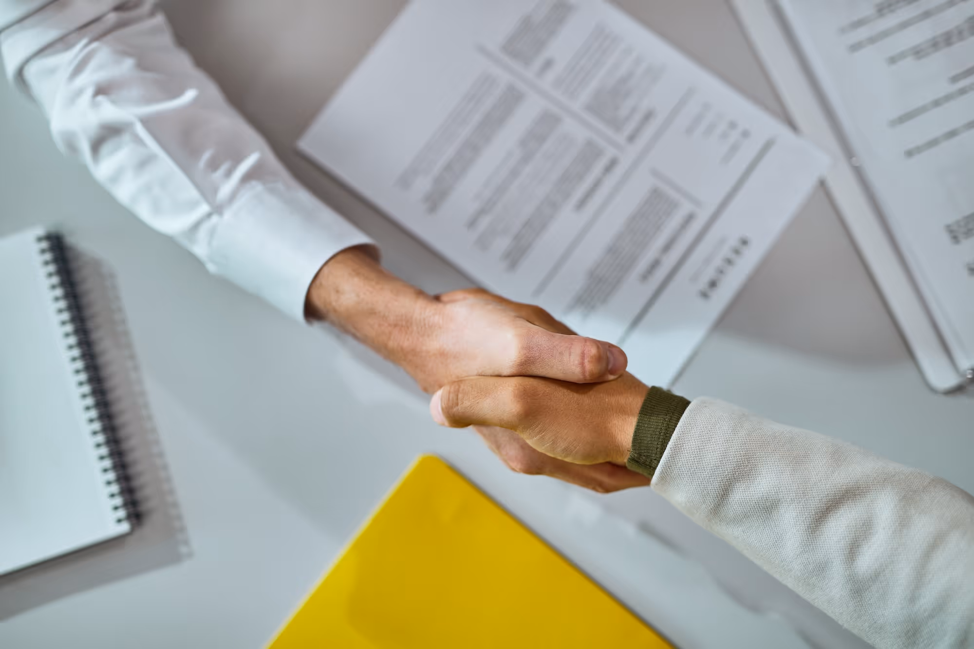 Two people shaking hands over a desk with documents and a yellow folder.