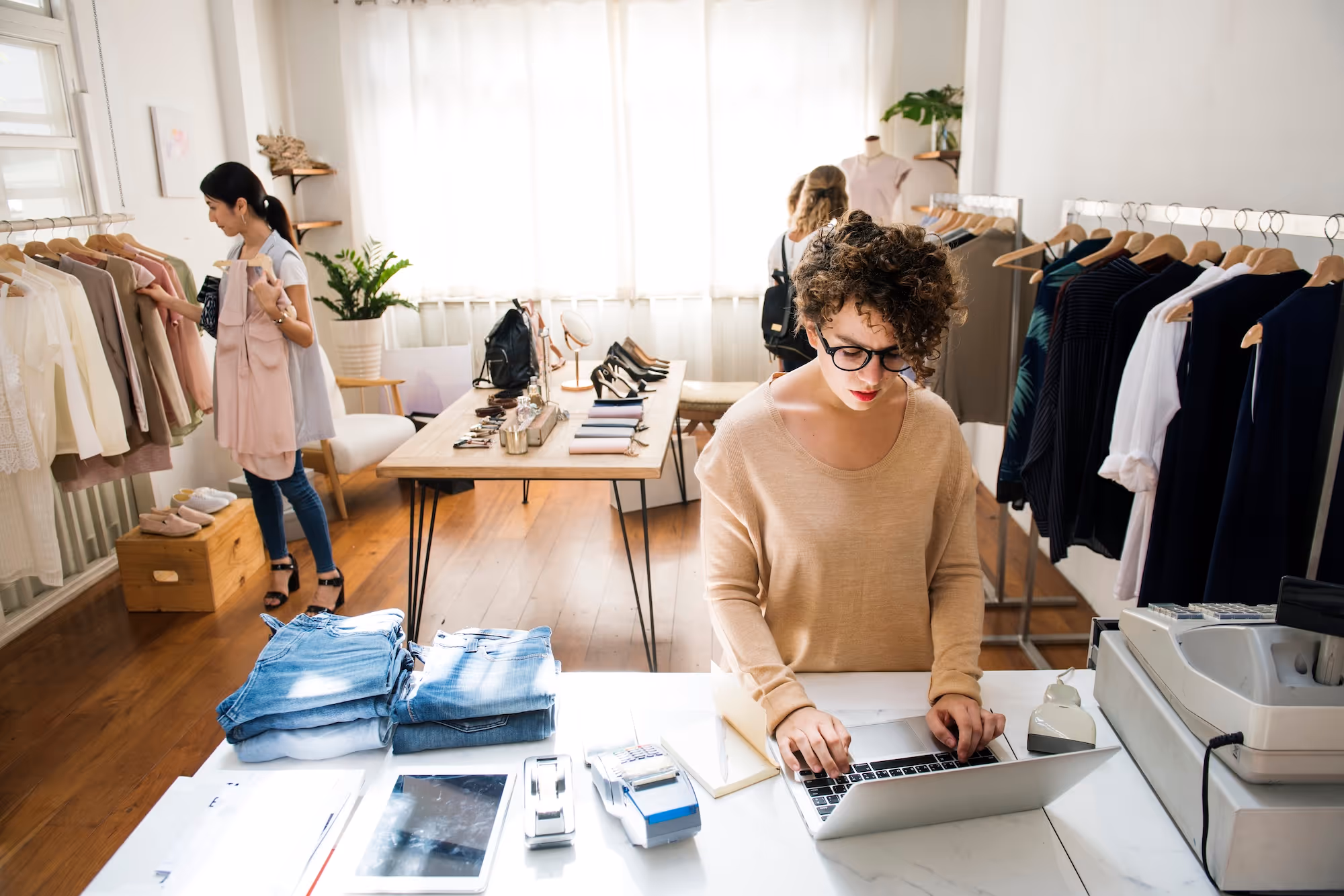 Woman with glasses working on a laptop at a clothing store counter with apparel and accessories around.