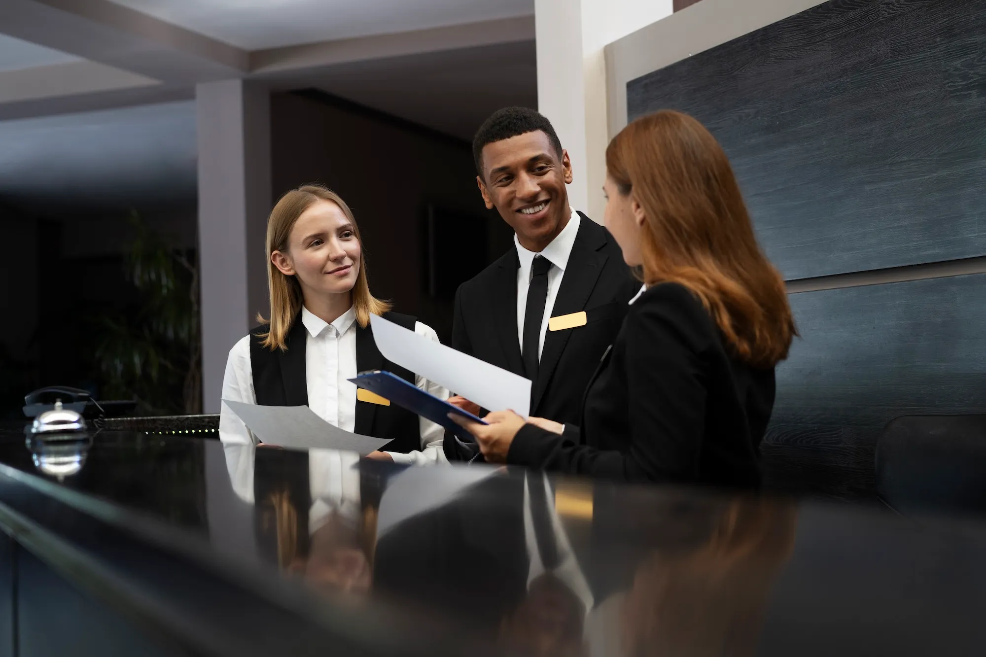 Three receptionists in elegant suits holding papers and talking behind a hotel front desk.