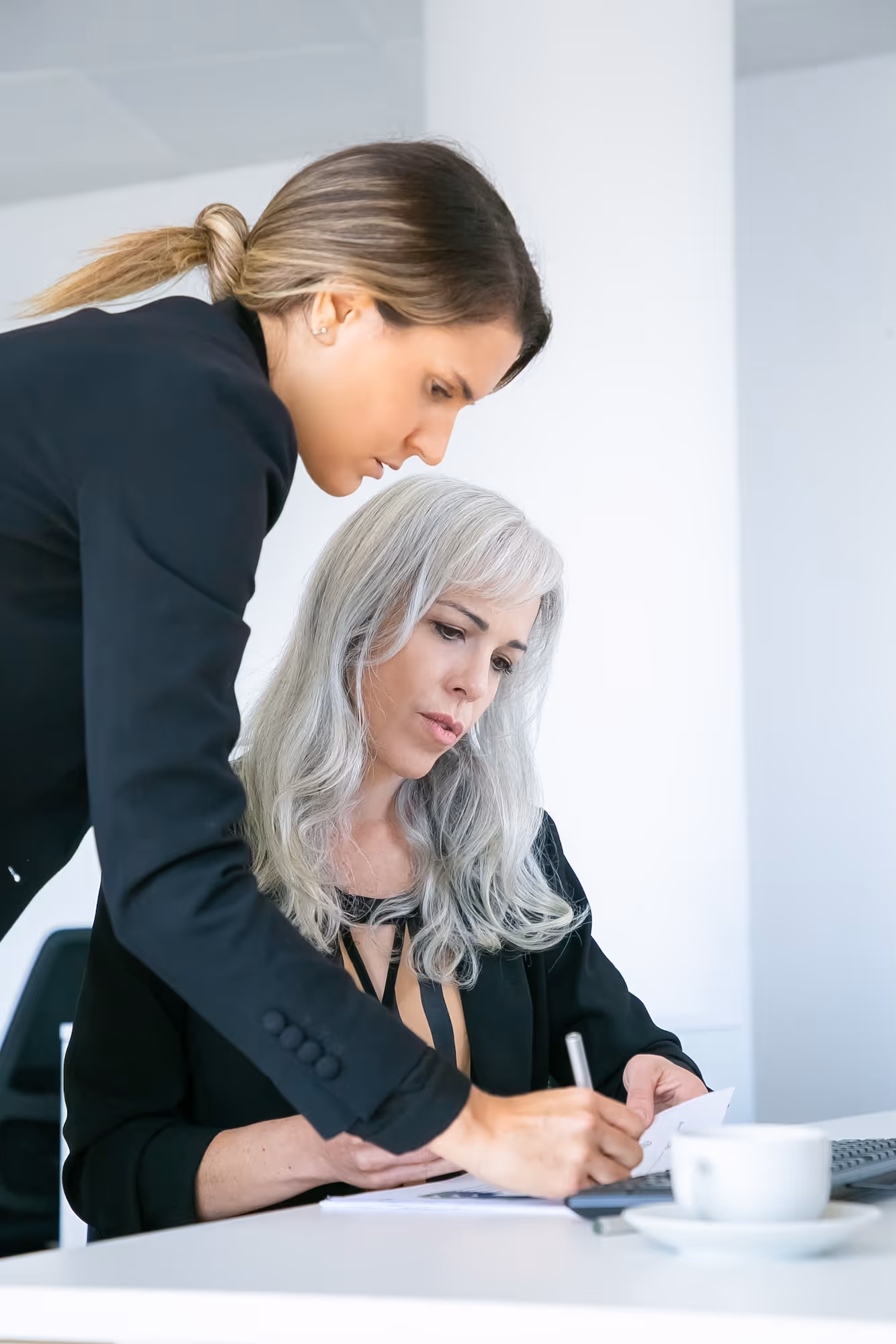 Two professional women collaborating at a desk, one writing notes while the other watches closely.