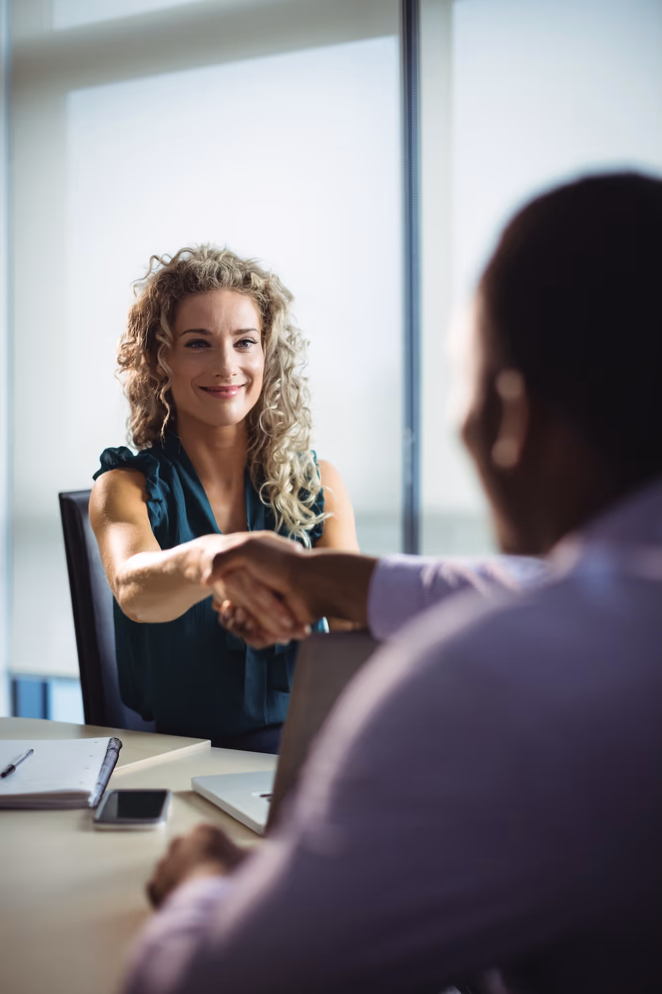 Two business professionals shaking hands across a desk in an office setting.