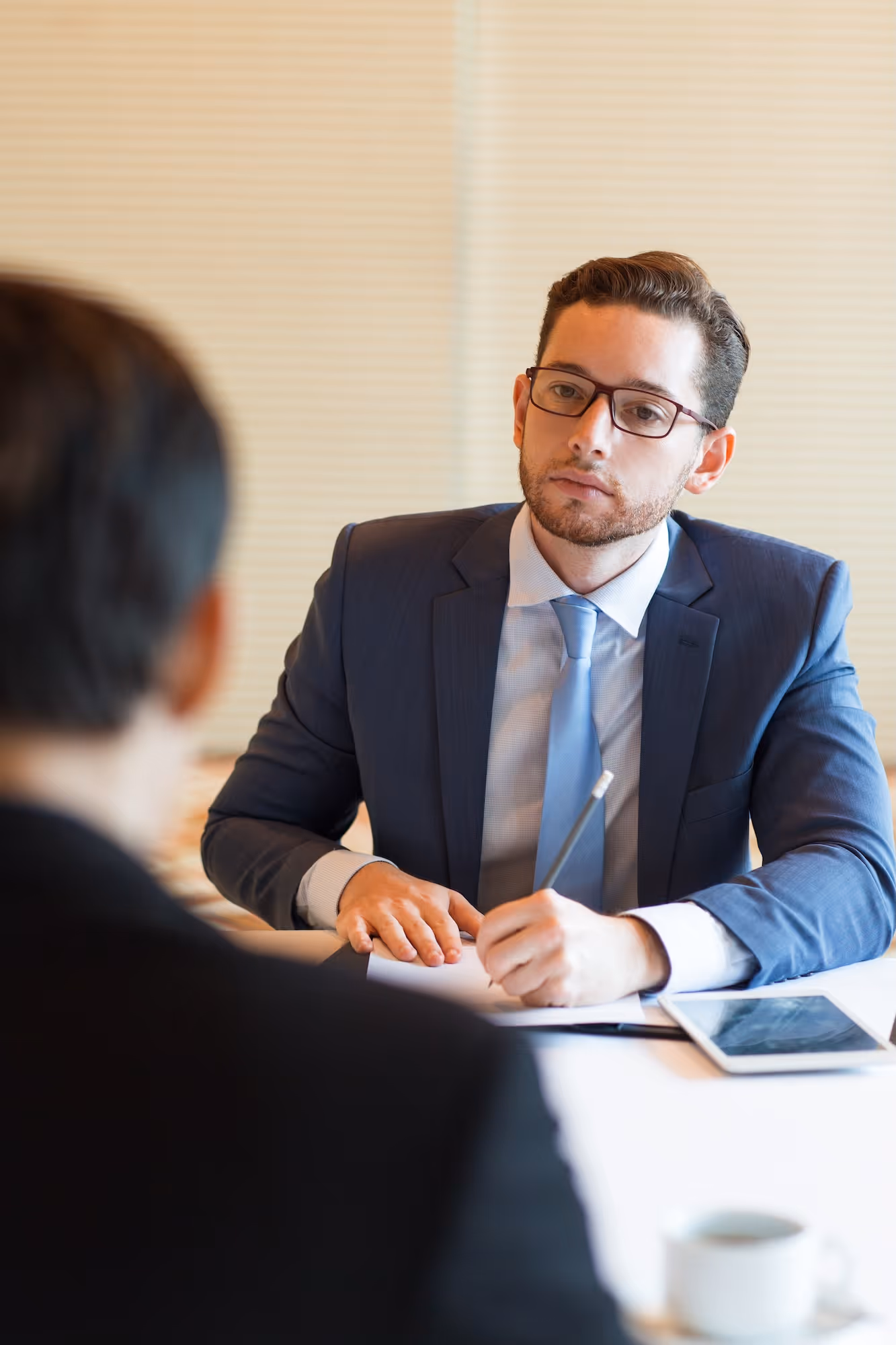 Man in glasses and suit attentively interviewing another person, taking notes with a pen.