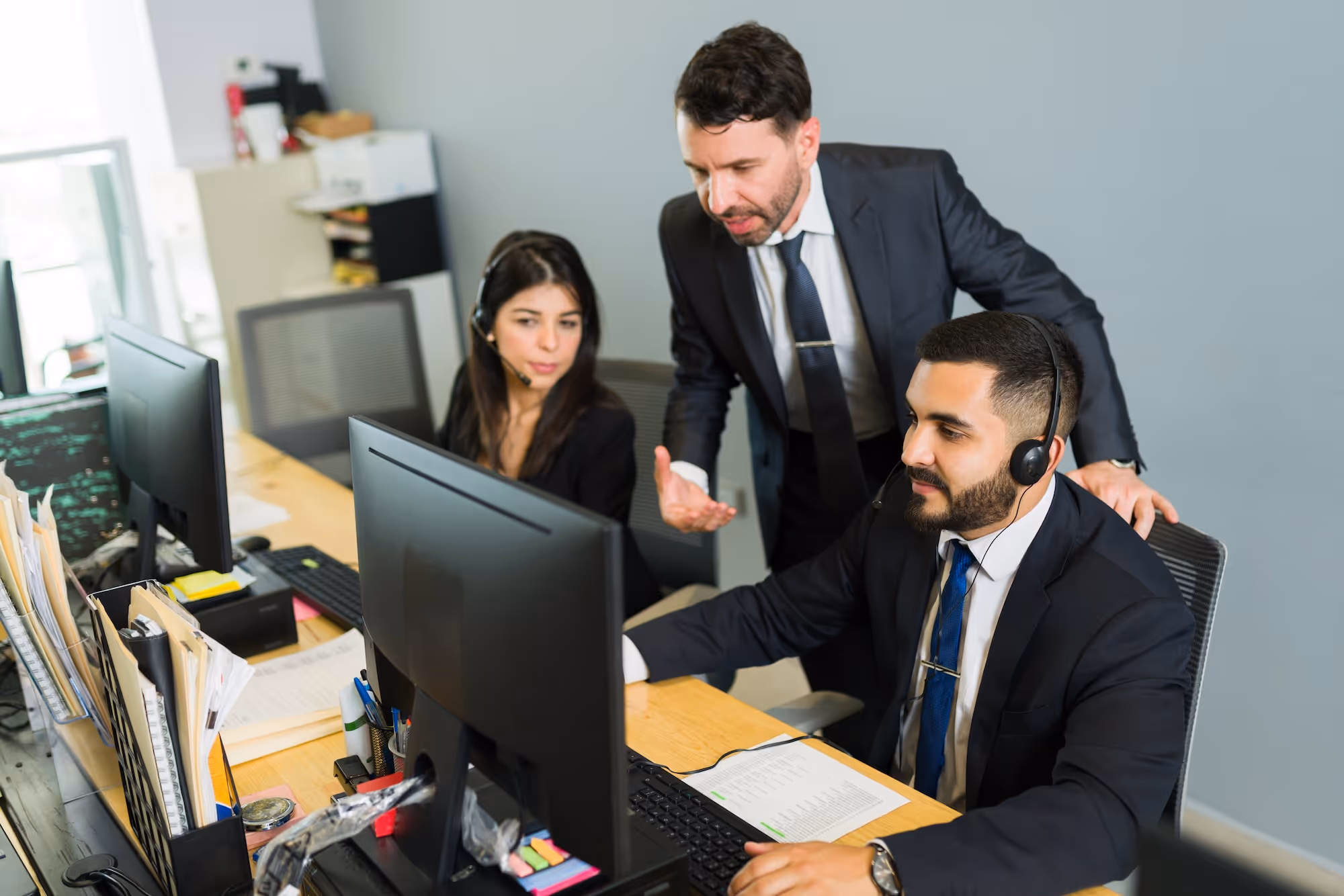 Male supervisor in suit guiding two call center agents wearing headsets at their desks.