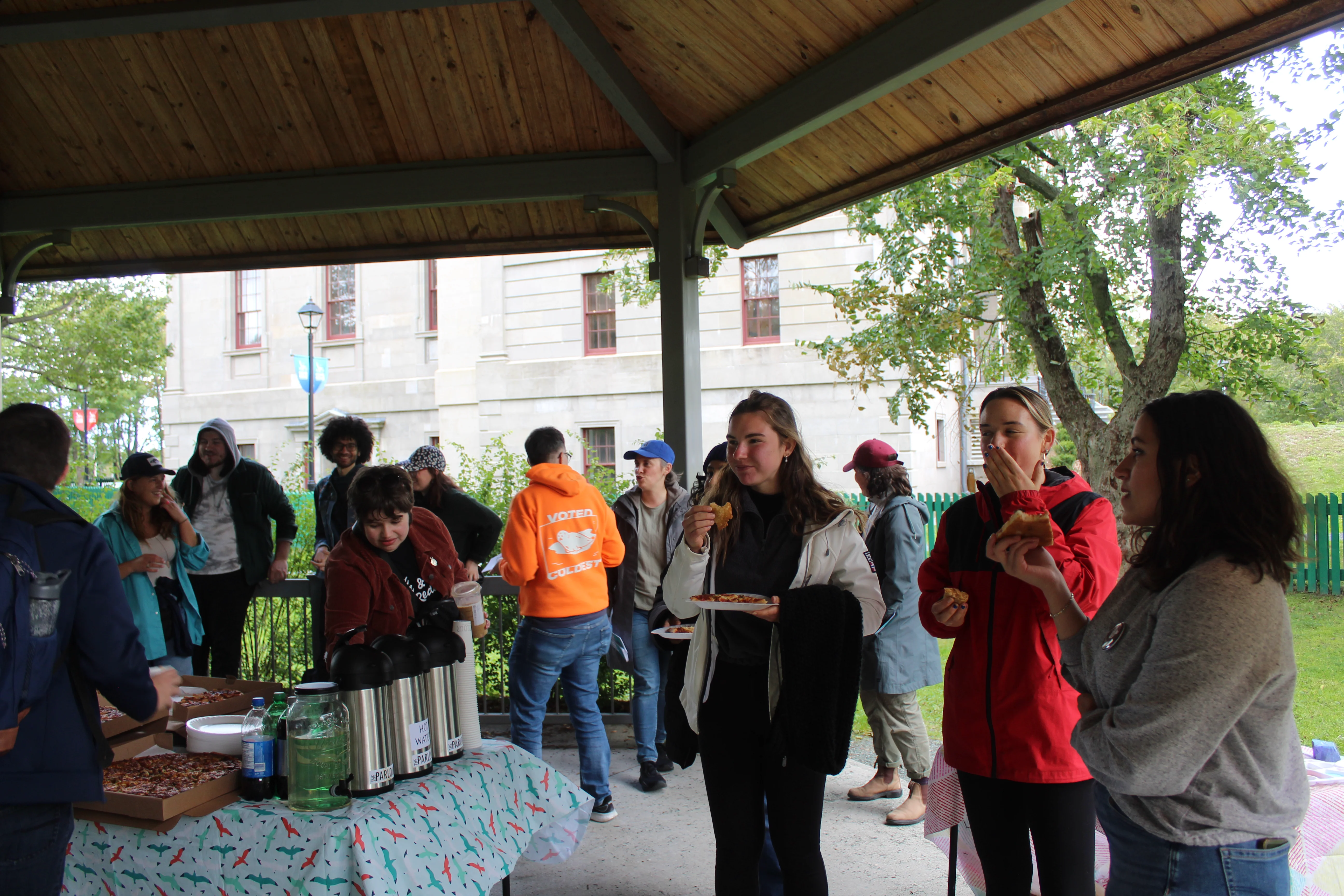 Community members sharing food and connecting at an outdoor MAC gathering under a wooden gazebo structure.