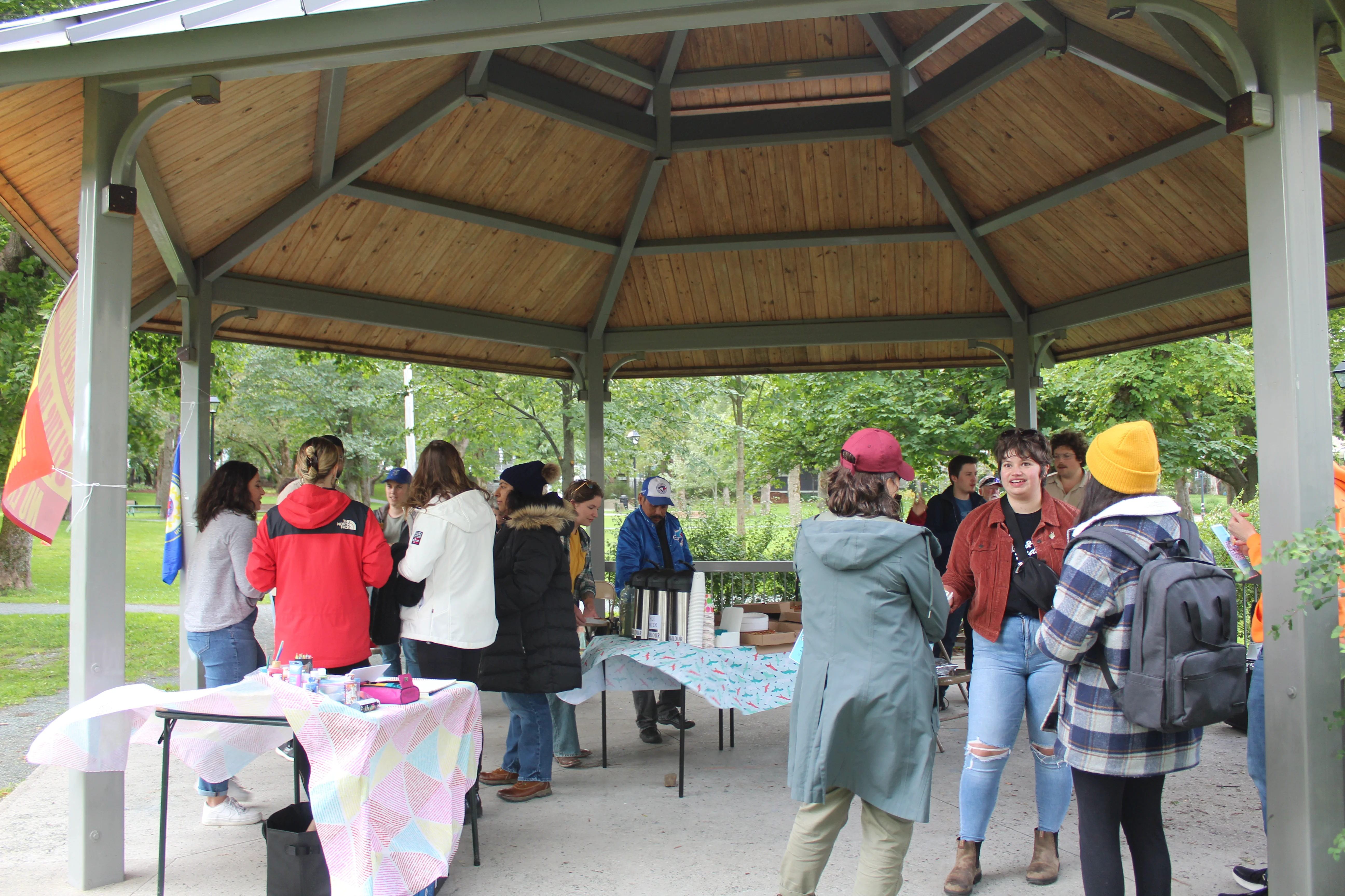 Group of people socializing and standing around tables under a wooden pavilion in a park.
