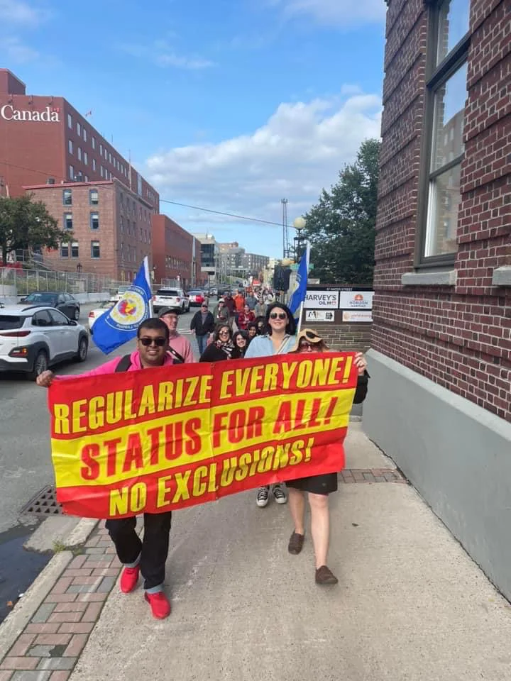 People marching in the street holding a red and yellow banner reading Regularize Everyone, Status for All, No Exclusions