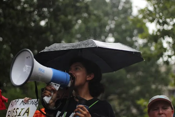 An organizer speaking into a megaphone at a rally in the rain, with community members visible around them.