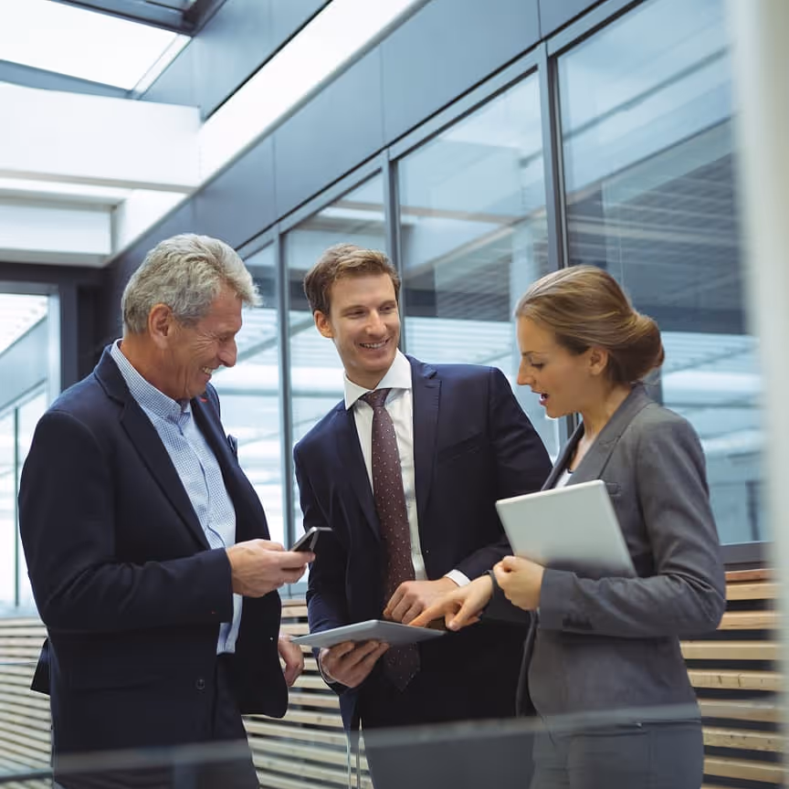 Three professionals in suits discussing work using tablet and phone in a modern office.