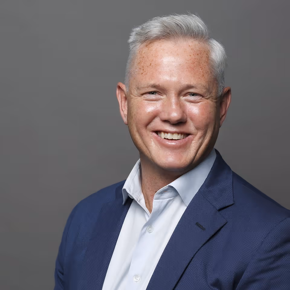 Smiling middle-aged man with gray hair wearing a blue suit and white shirt against gray background.