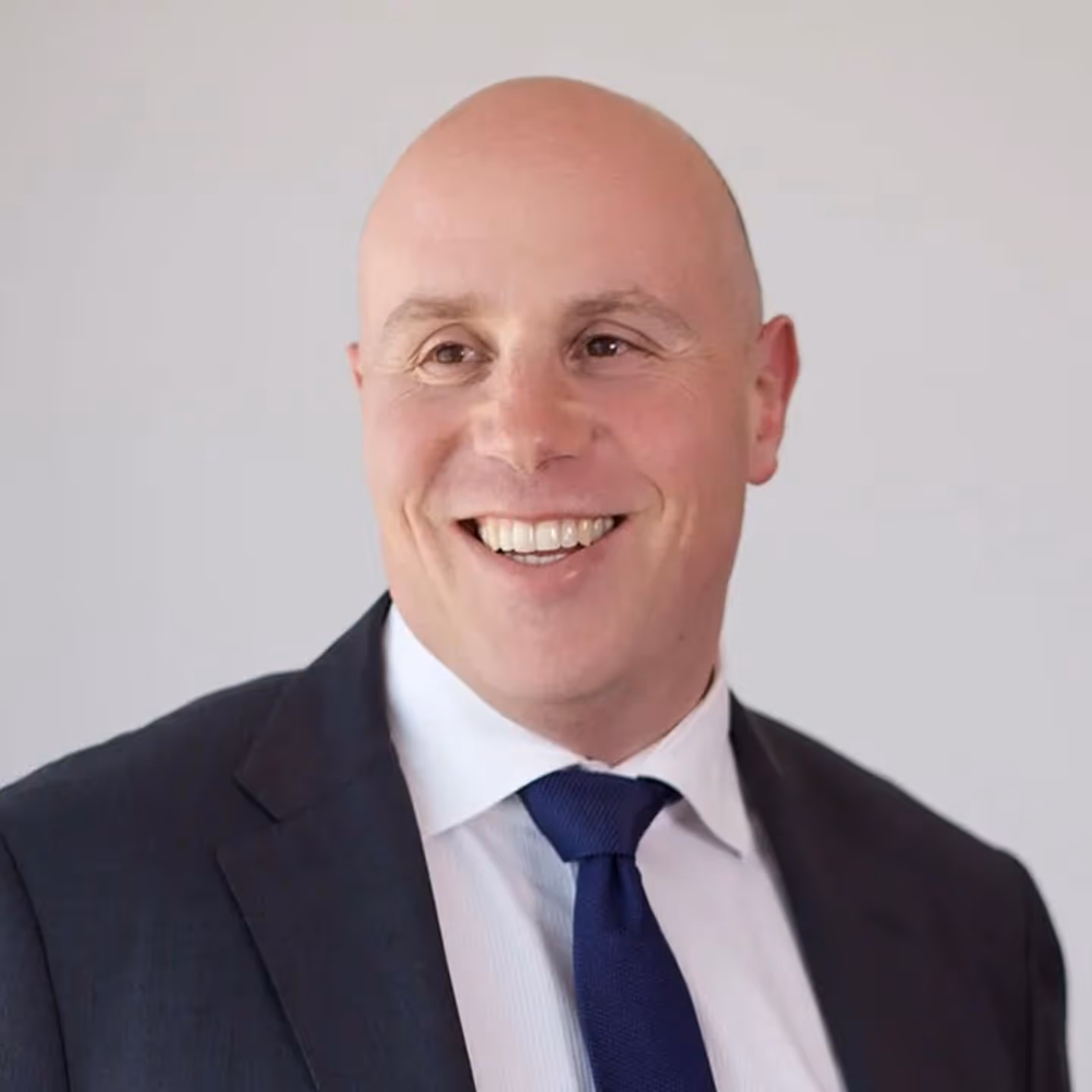 Smiling wearing a dark suit, white shirt, and blue tie against a plain background.