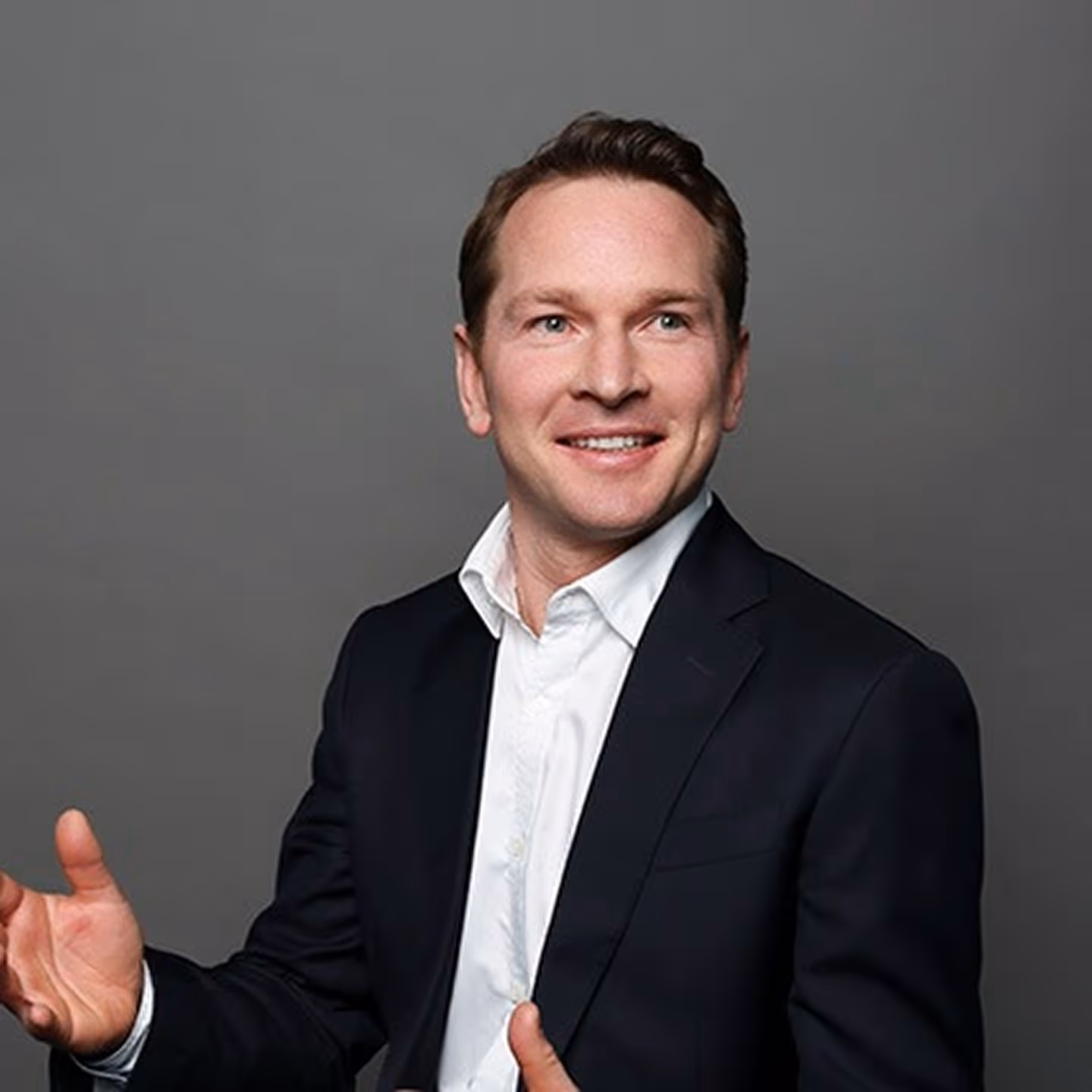 Smiling man in a dark suit jacket and white shirt gesturing with hands against a gray background.
