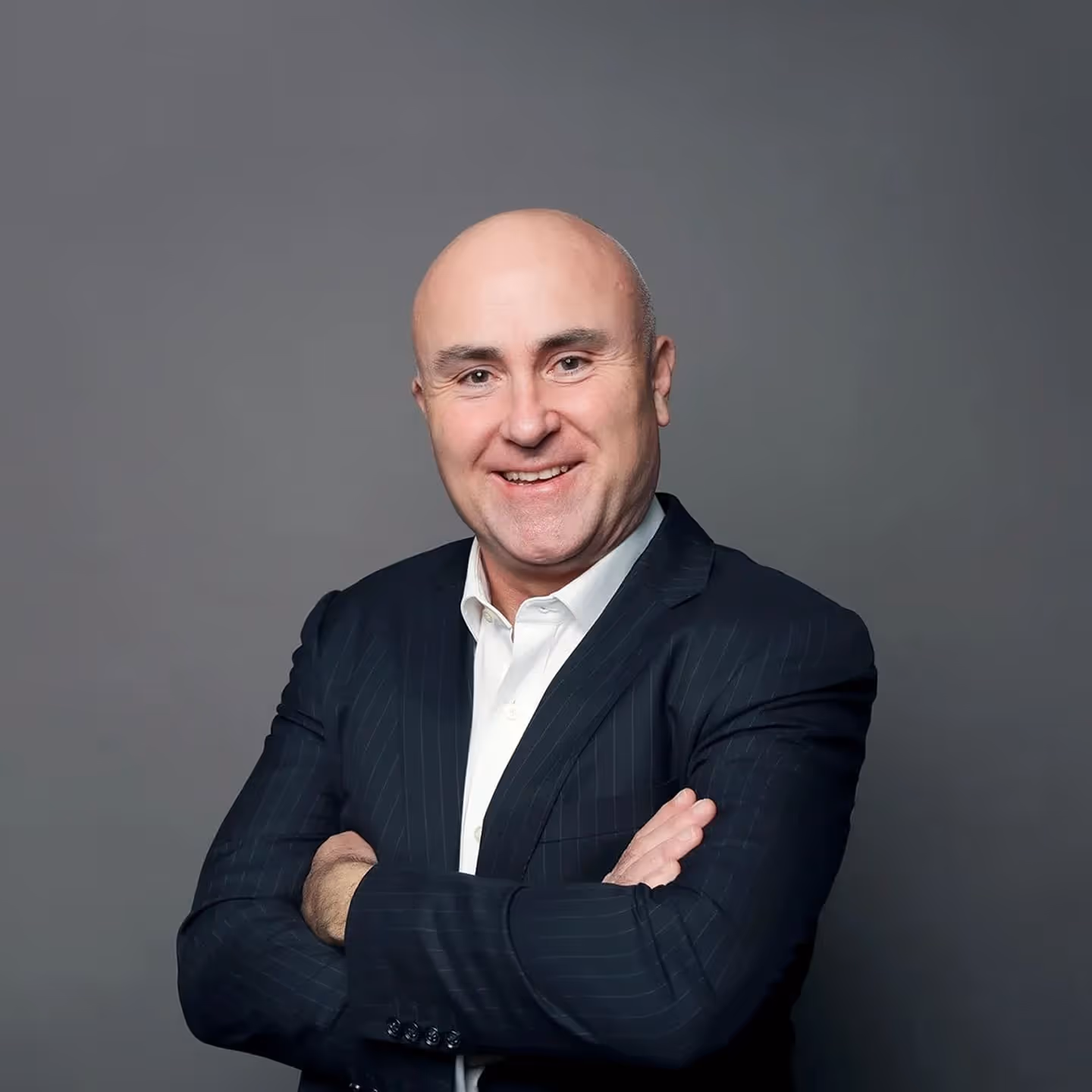 Smiling bald man in a dark pinstripe suit with white shirt crosses arms against gray background.