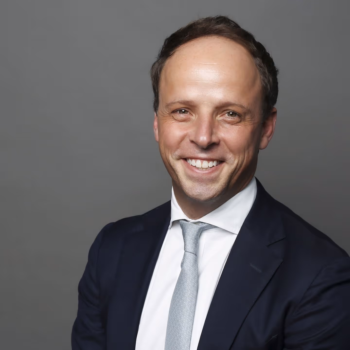 Smiling man in dark suit, white shirt, and light gray tie posing against a plain gray background.