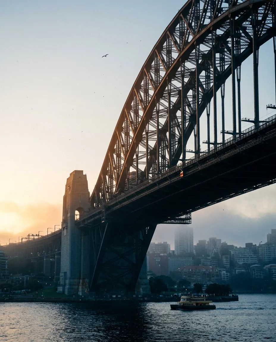Sydney Harbour Bridge arch with city skyline, water, a ferry, and a bird at sunrise or sunset.