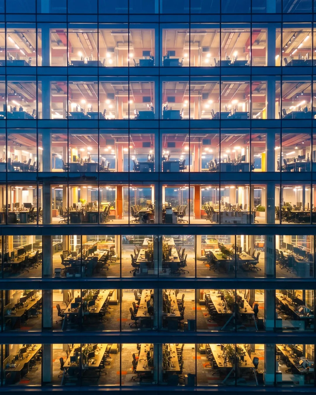Exterior view of a multi-story office building with rows of desks and chairs lit up at night.