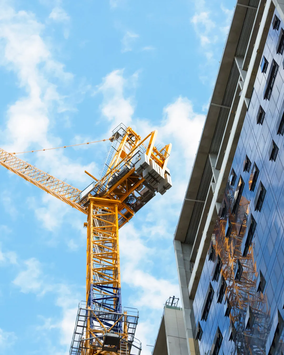 Yellow construction crane beside a modern glass building under a partly cloudy blue sky.