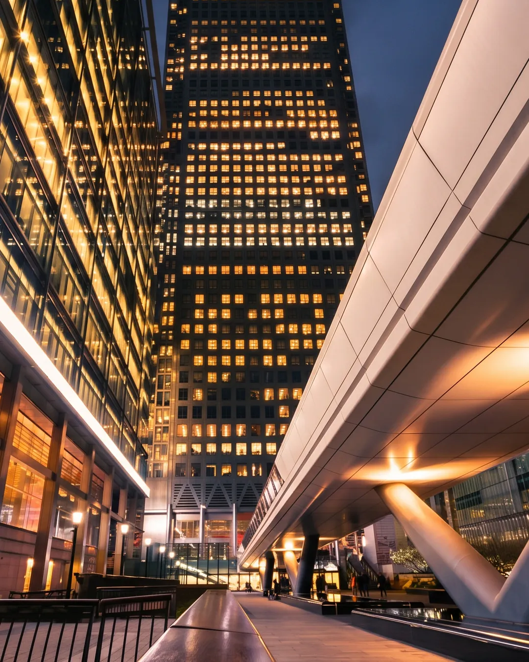 Night view of illuminated modern skyscrapers and a futuristic pedestrian bridge in a city plaza.