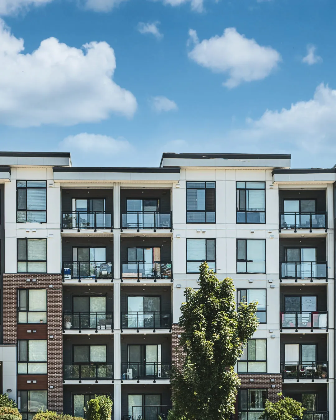 Modern apartment building with balconies against a blue sky with scattered clouds and trees in front.
