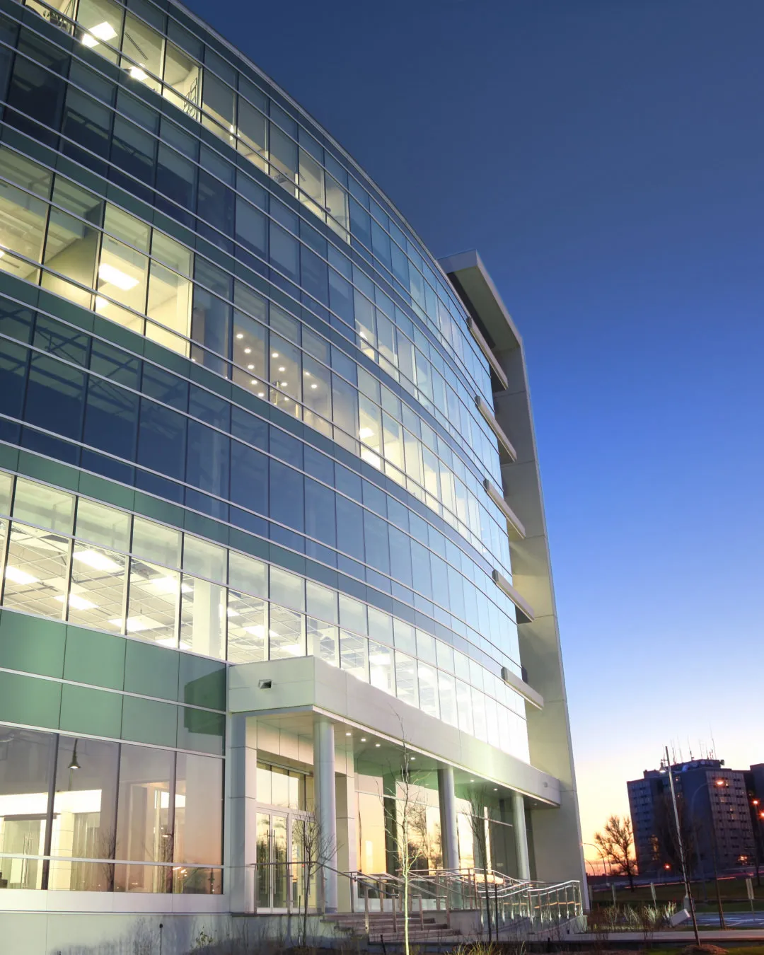Modern glass office building curved facade lit from inside at dusk with clear sky background.