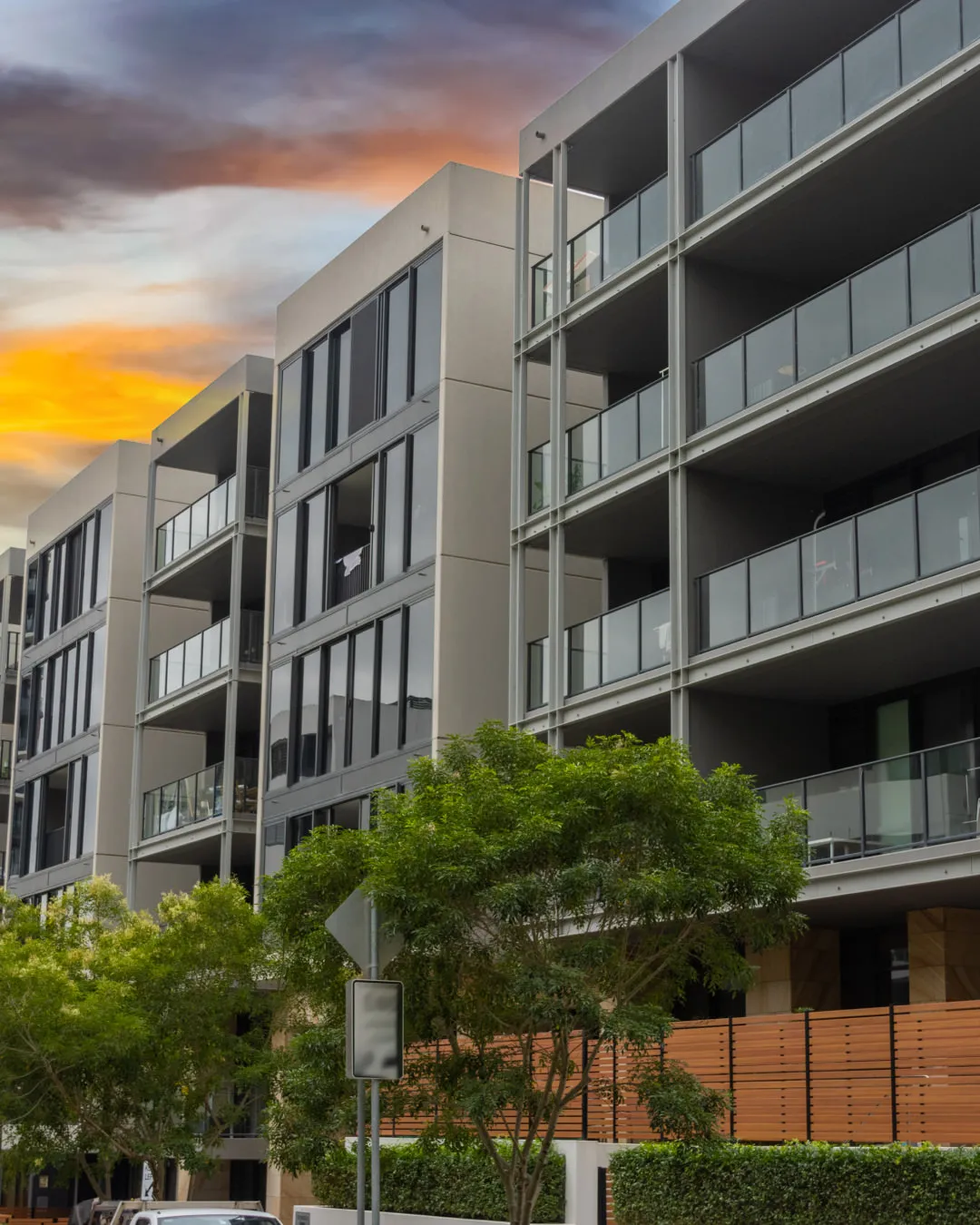 Modern apartment buildings with glass balconies and trees in front at sunset.