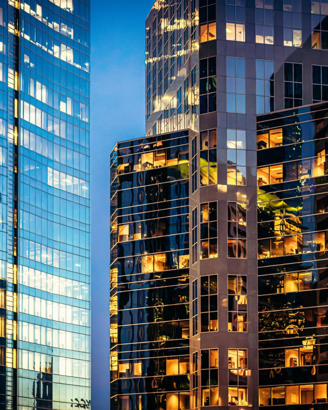 Two nearby modern office buildings with many lit windows reflecting the evening sky and city lights.