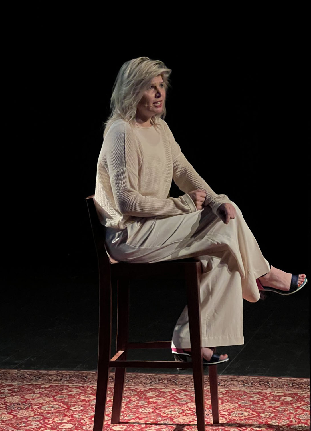Woman in beige sweater and pants sitting cross-legged on a tall wooden chair on a patterned rug against a dark background.