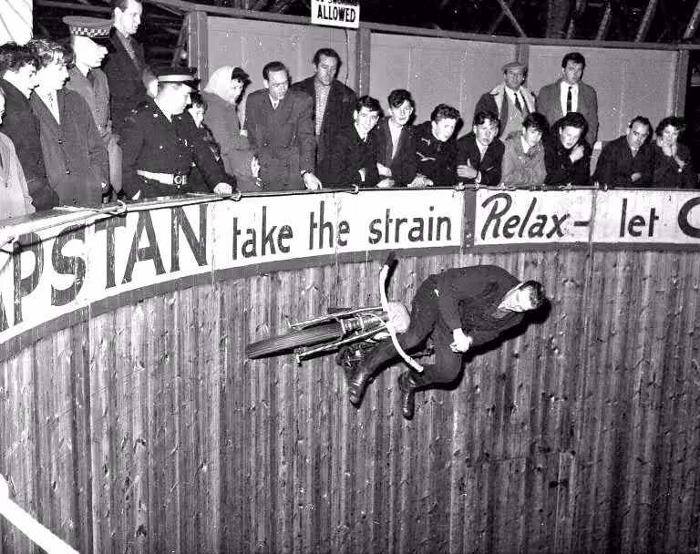 Zwart-wit foto van een motorcoureur die horizontaal rijst op een houten muur in een Wall of Death-show, terwijl een menigte van toeschouwers toekijkt vanaf de rand bovenaan.