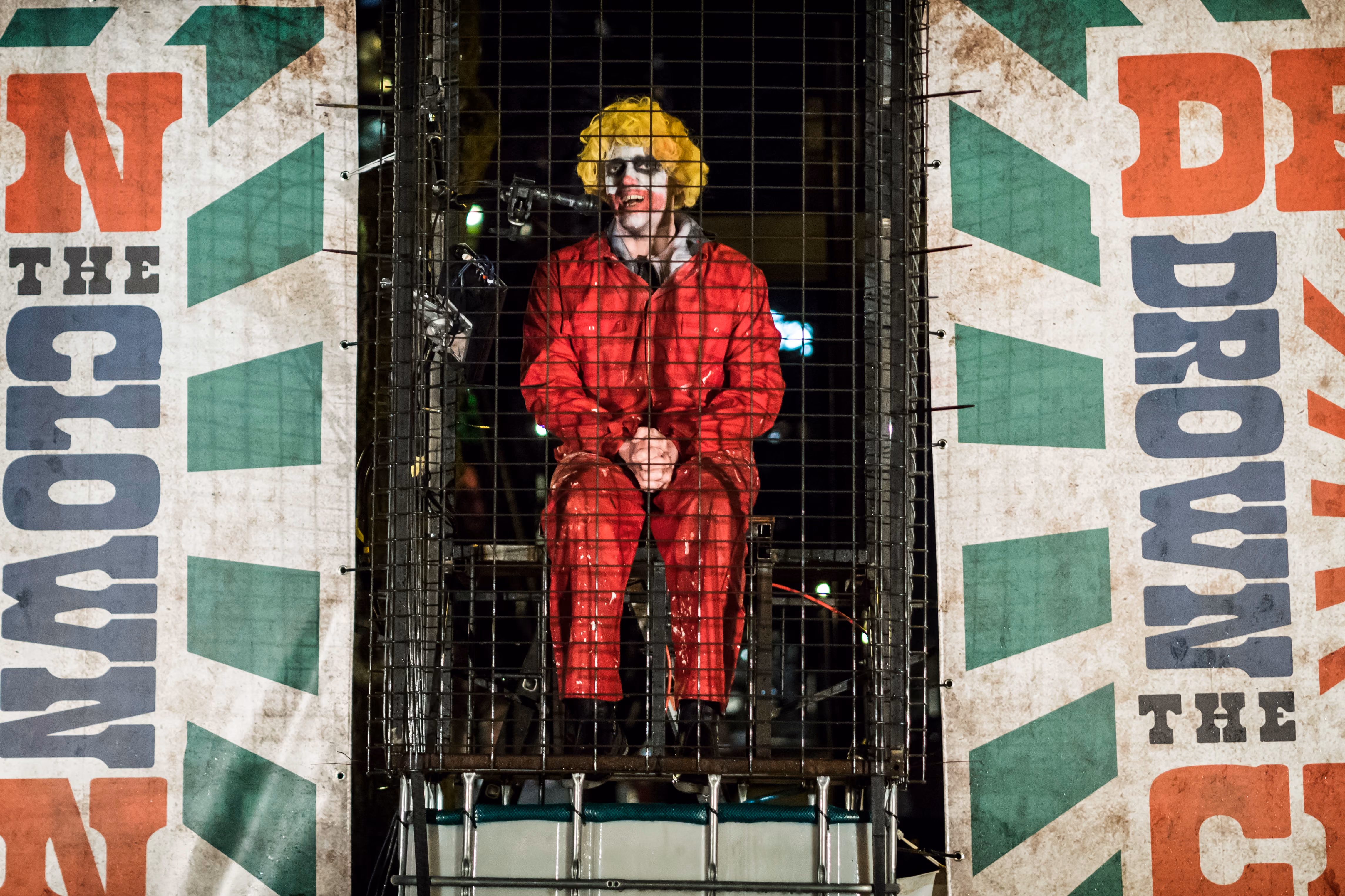 Man met clownsmake-up, wit geschminkt gezicht en rode lippen, een gele pruik en een rood jasje met rode broek, zittend in een kooi die is klaargemaakt voor een dunk tank tijdens een evenement in de avond. Rondom de kooi staat een bord met de tekst ‘Dunk the Clown’ in grote rode, zwarte en blauwe letters, tegen een groen-witte achtergrond met zonnestralen.