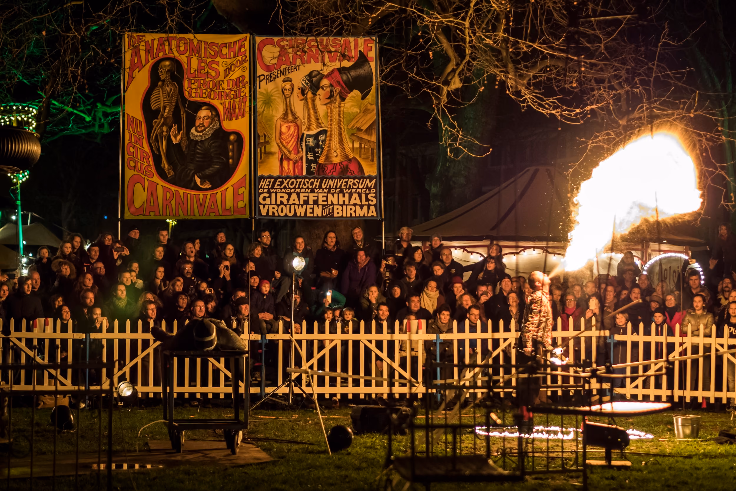 Een vuurspuwer voert een vuurshow uit bij nacht voor een grote menigte achter een houten hek bij een carnavalsfeest met kleurrijke posters aan palen.