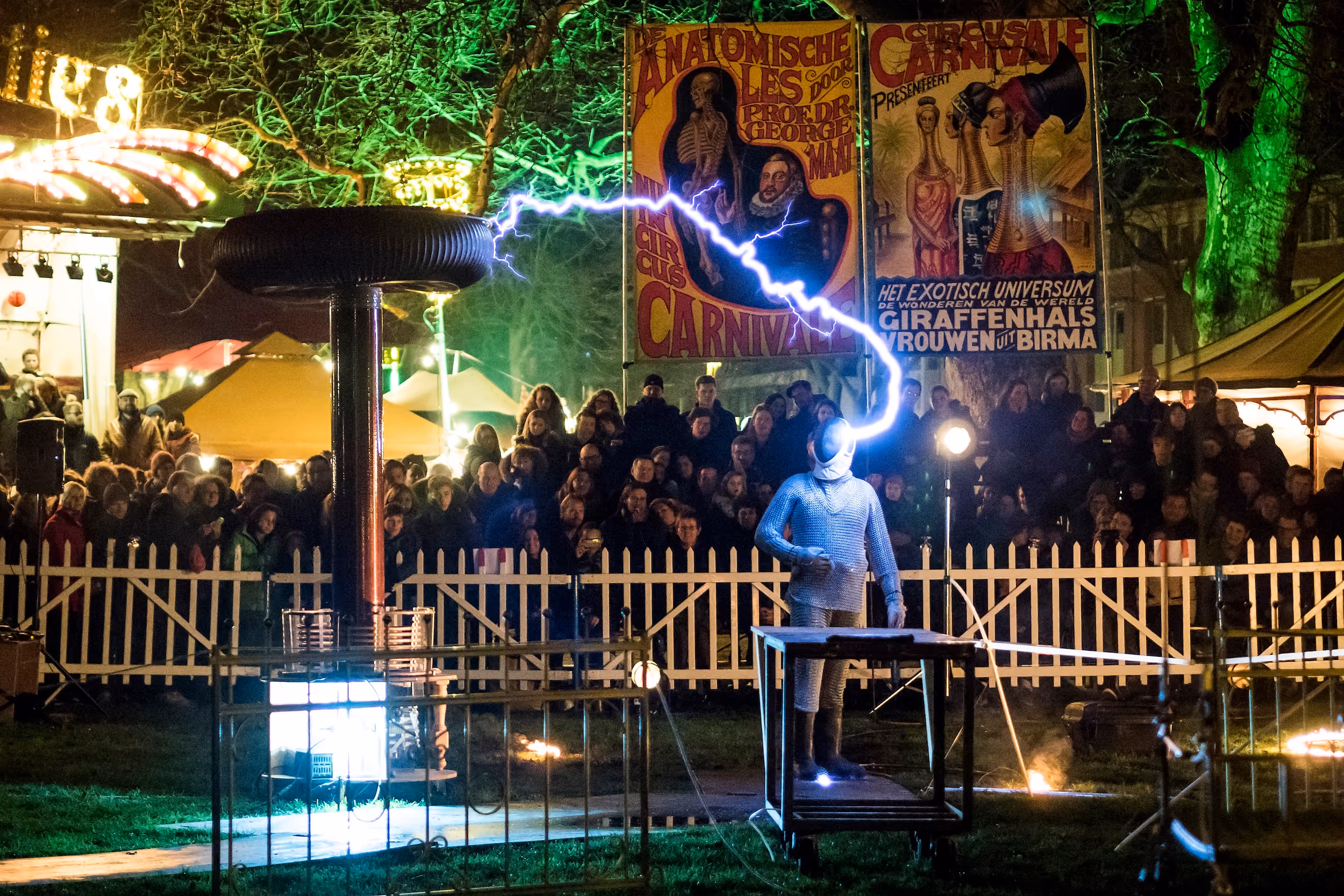 Een persoon in een metalen harnas staat buiten 's nachts voor een publiek en wordt getroffen door een bliksemschicht van een Tesla-spoel naast hen, met kleurrijke festivalposters en bomen op de achtergrond.