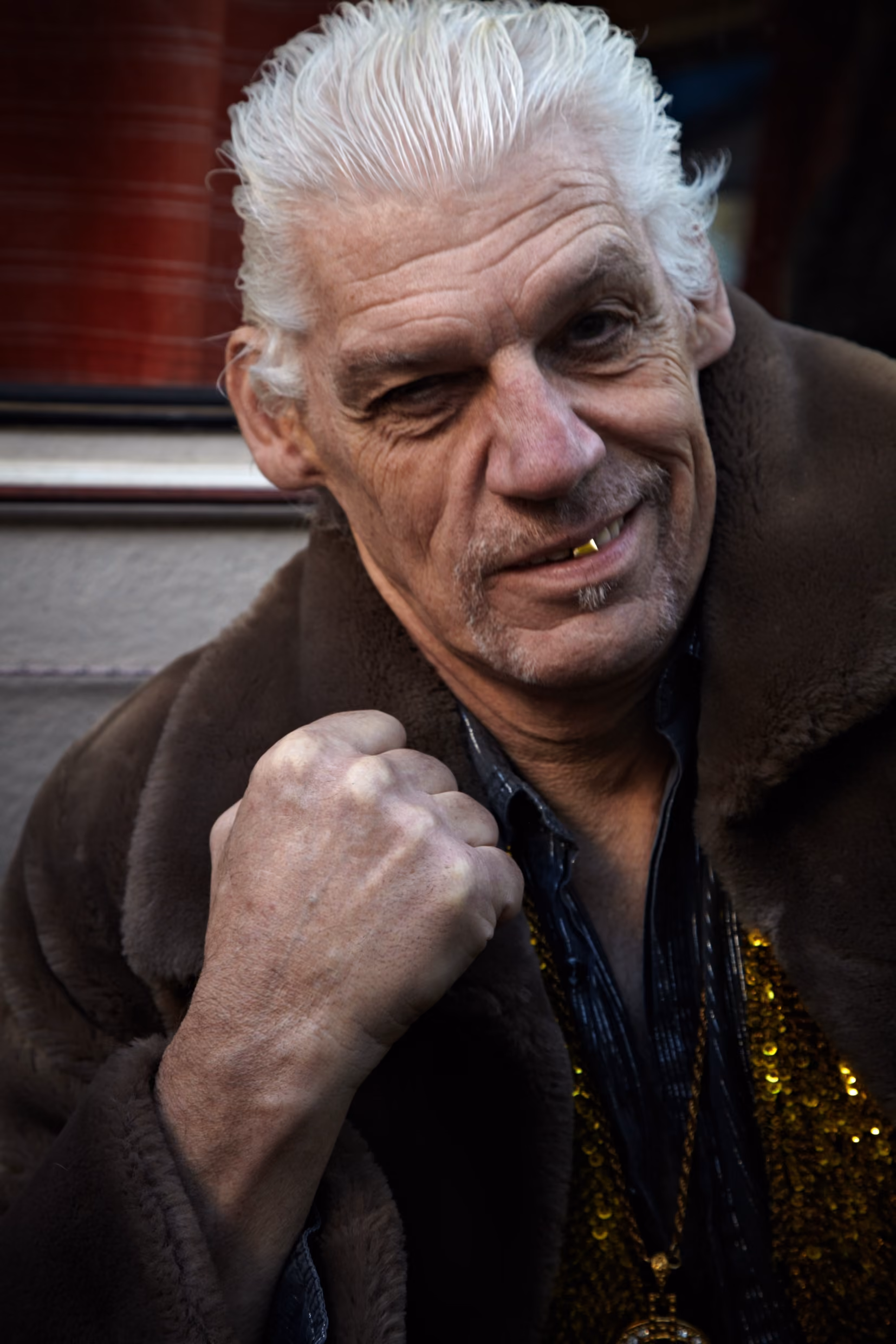 Fritz Parlando: een oudere man met wit haar draagt een bruine jas en een goudkleurige ketting, kijkt naar de camera met een gefronste blik en een vuist omhoog.