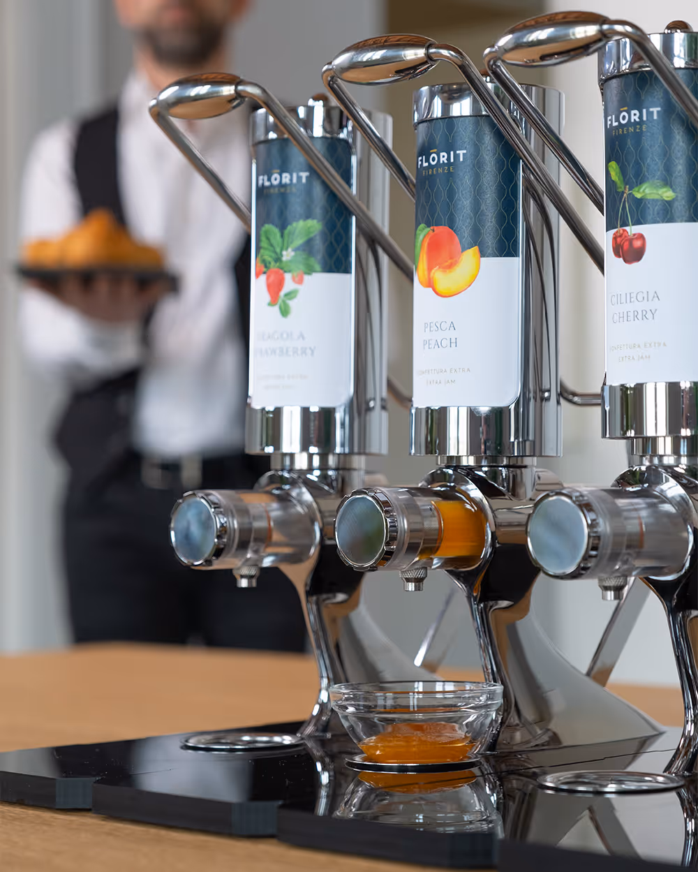 Three stainless steel jam dispensers labeled strawberry, peach, and cherry with a waiter in the background holding a plate of croissants.