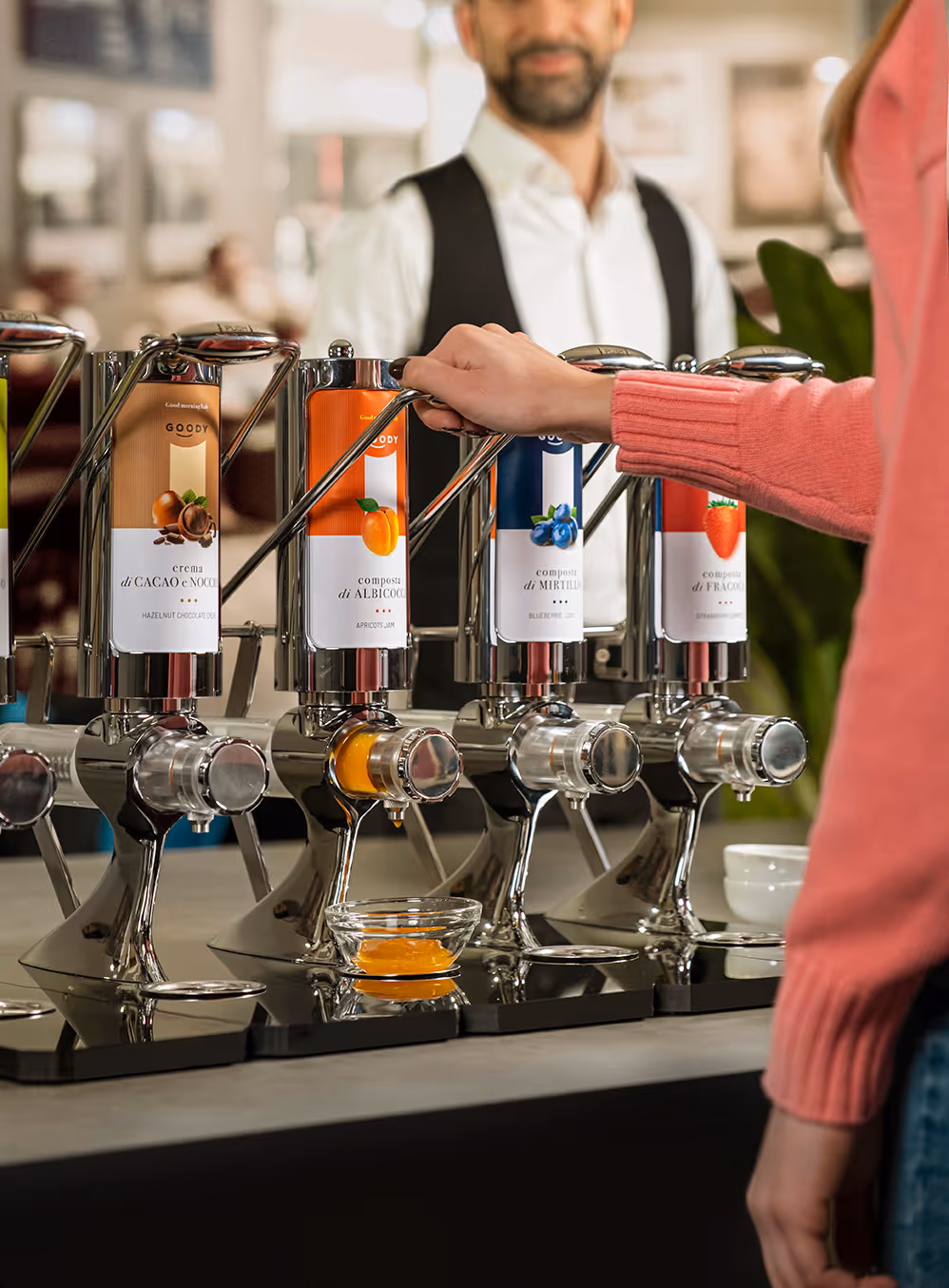 Person dispensing apricot jam from a stainless steel dispenser with other dispensers labeled hazelnut chocolate cream, blueberry jam, and strawberry jam visible.