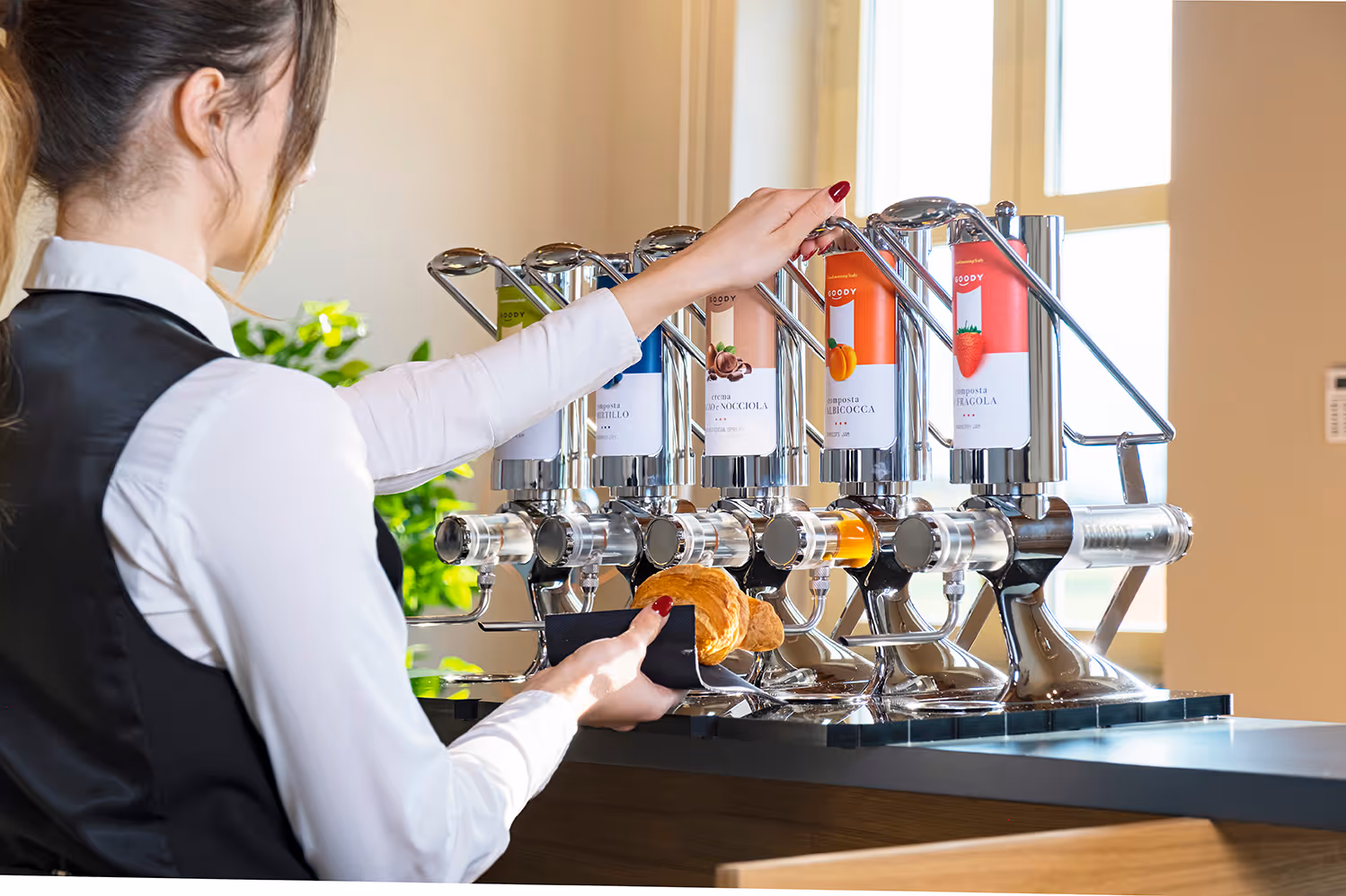 Woman using a multi-flavor spread dispenser to put spread on a croissant in a well-lit kitchen or cafe setting.