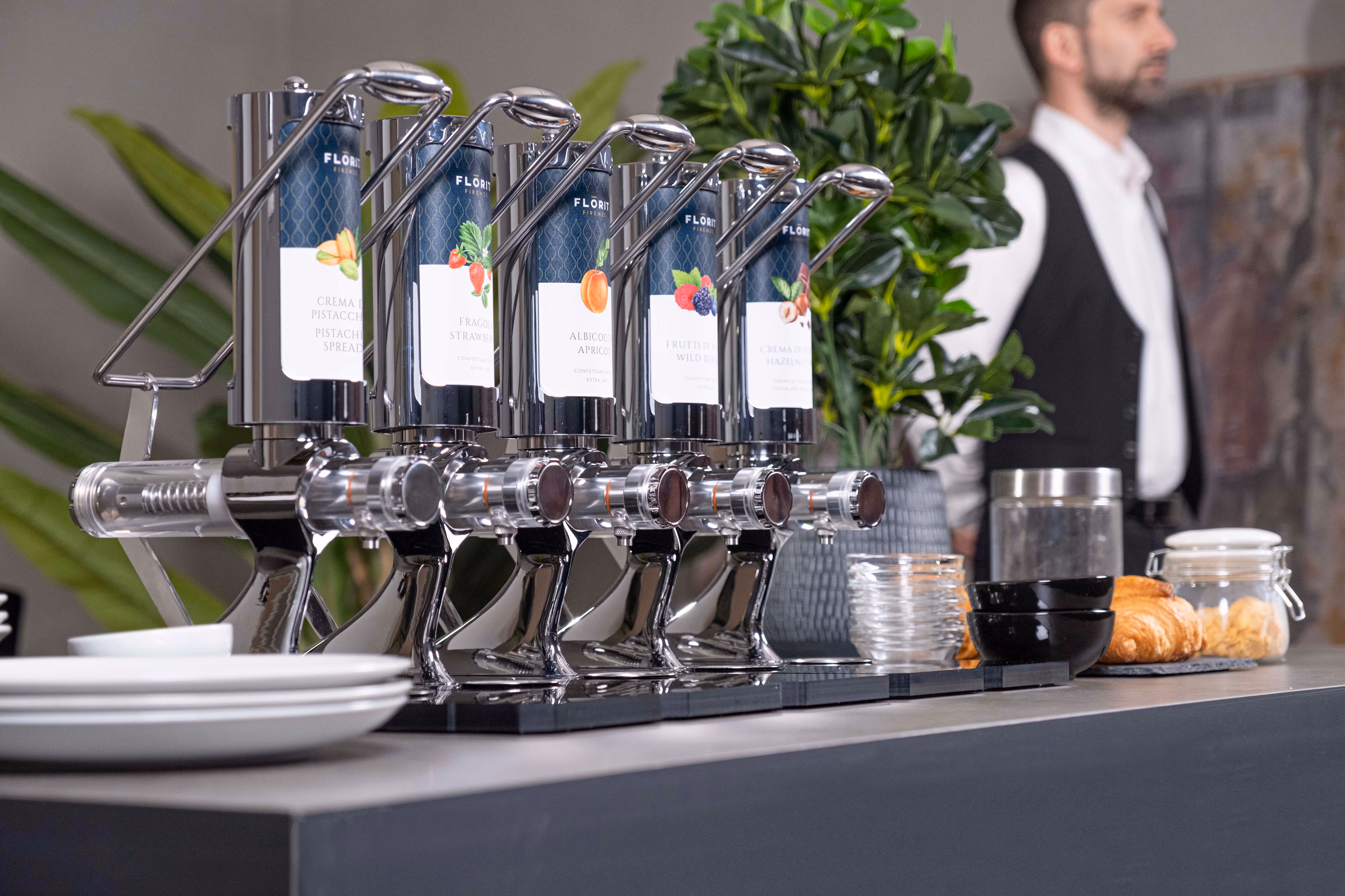 Row of six shiny dispensers labeled with different fruit spreads including pistachio, strawberry, apricot, wild berry, and hazelnut on a countertop with plates, bowls, croissants, and a blurred man in a vest in the background.