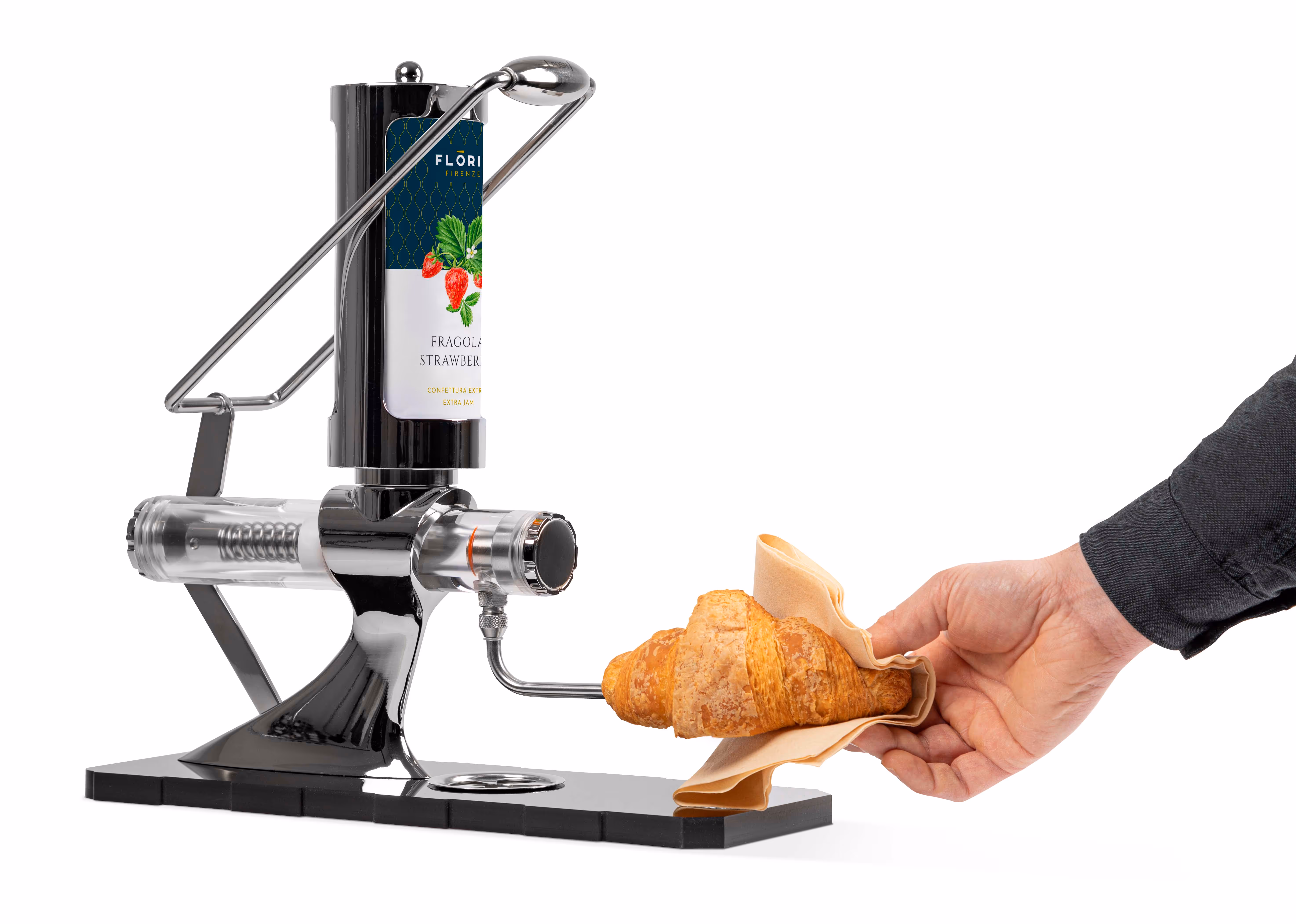 A hand holding a croissant wrapped in a napkin under a jam dispenser labeled strawberry extra jam.