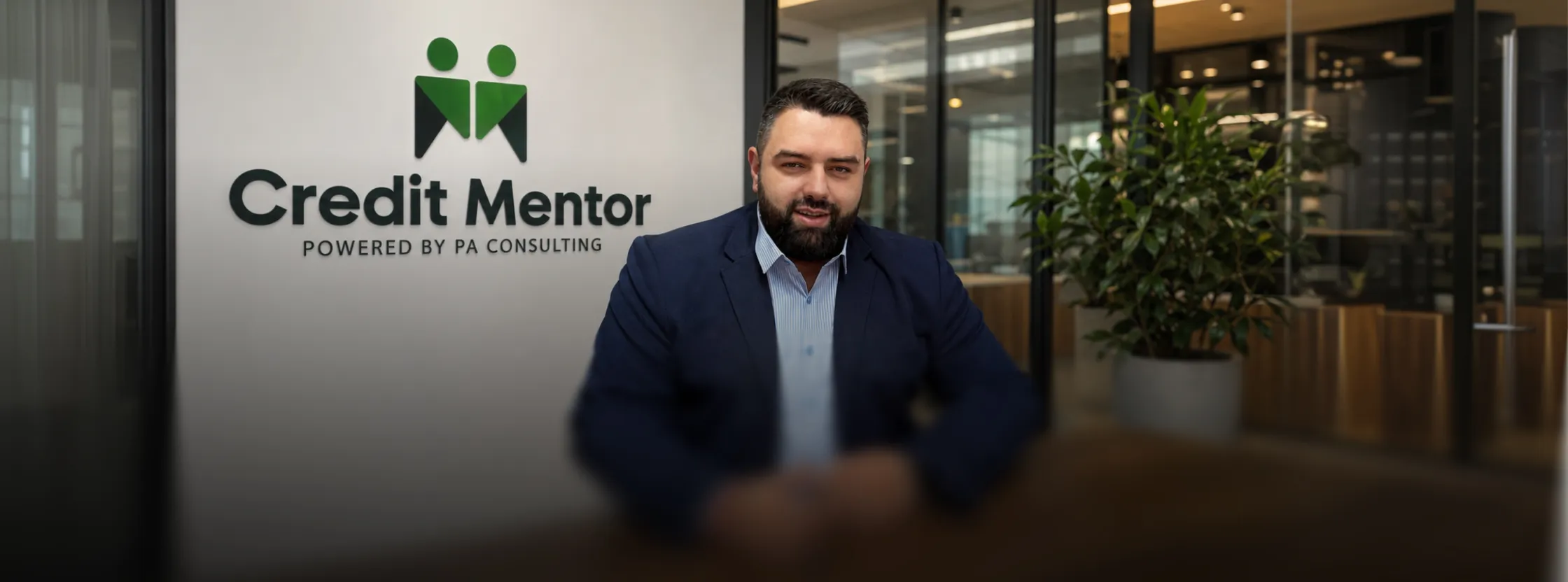 Мъж в синьо сако седнал пред стена с логото на Credit Mentor в офис.