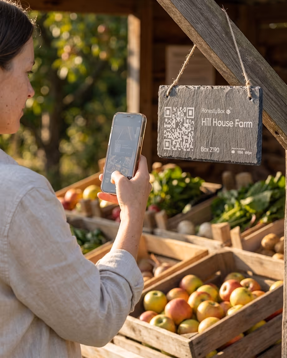 Person scanning a QR code sign for Hill House Farm at a farm stand with baskets of apples and vegetables.