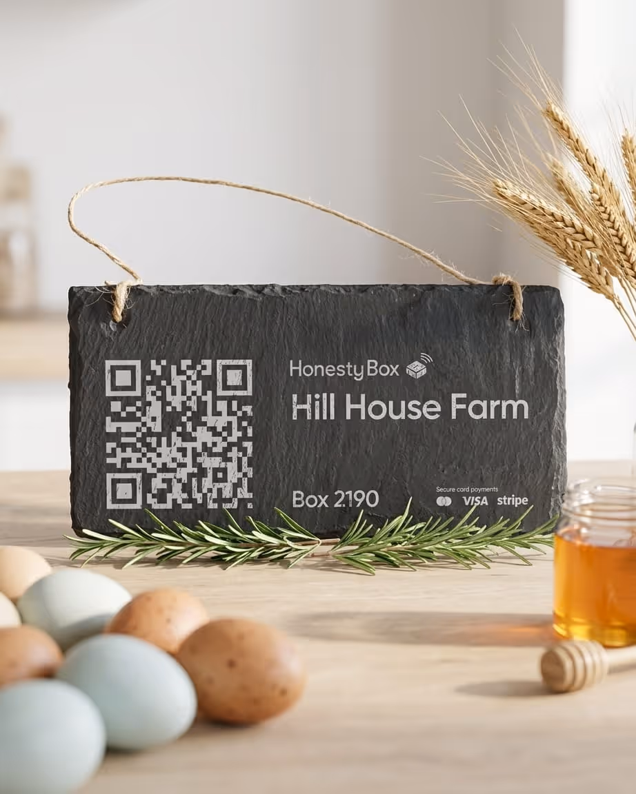 Slate sign with QR code and text 'HonestyBox Hill House Farm Box 2190', surrounded by eggs, sprigs of rosemary, wheat stalks, and a jar of honey with a honey dipper.