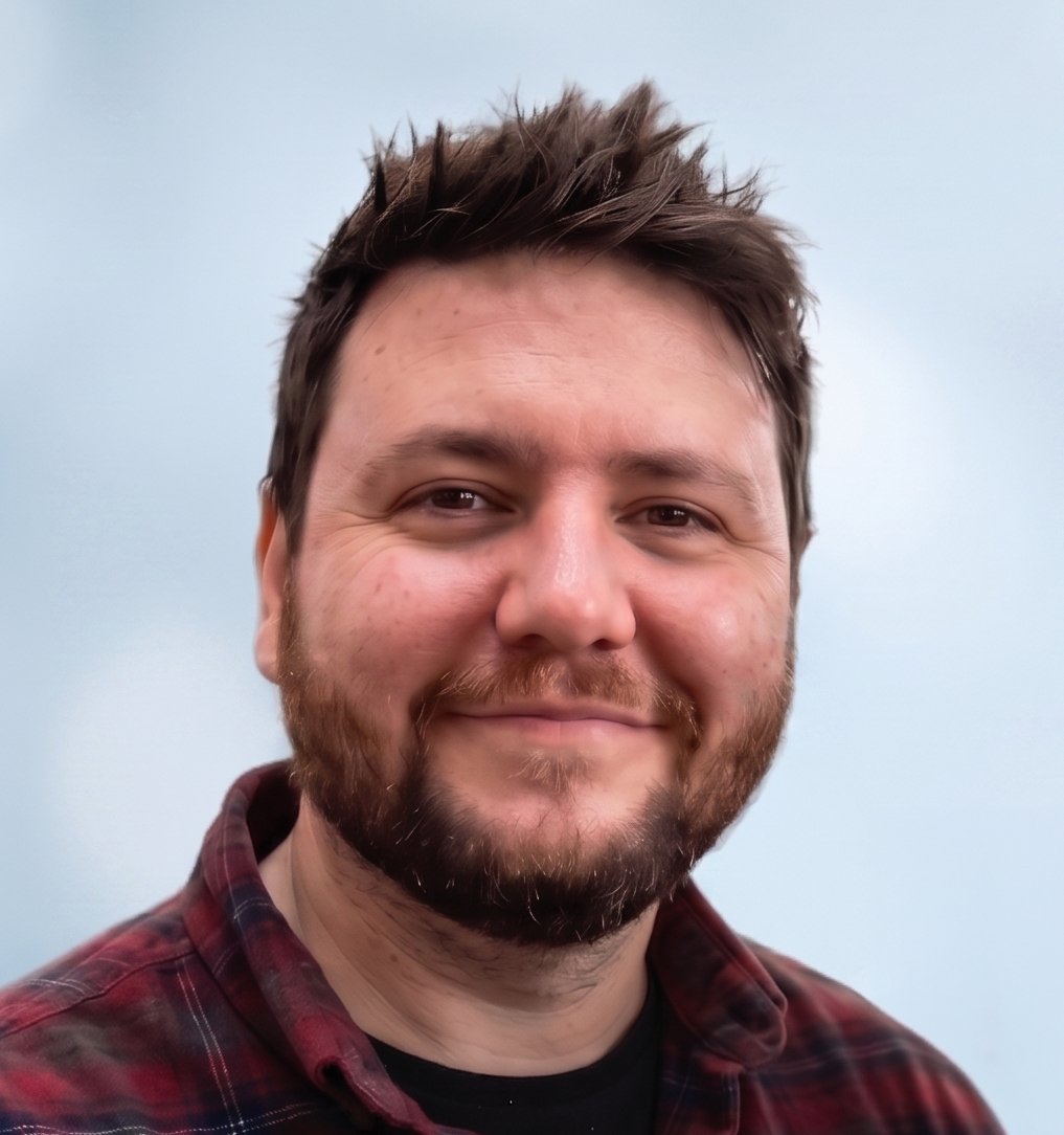 Smiling bearded man with tousled hair wearing a red and black plaid shirt against a light blue background.